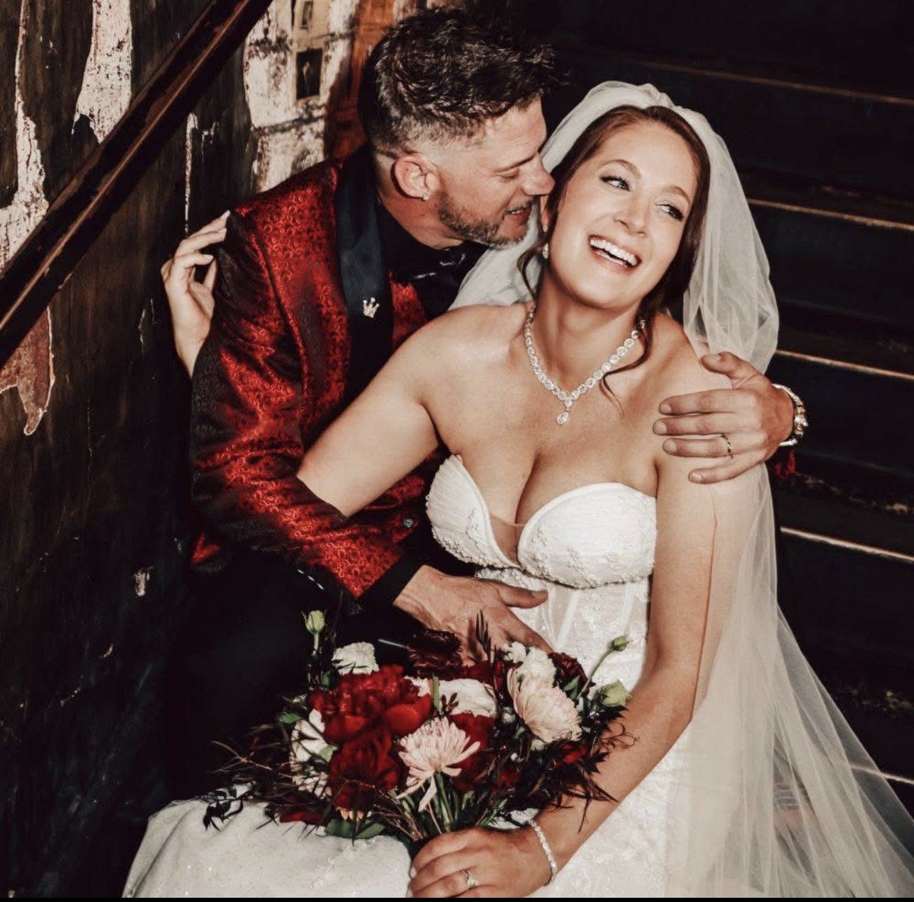 A bride and groom on their wedding day, smiling and embracing. The bride is holding a bouquet of red and pink flowers, wearing a white strapless wedding gown and a pearl necklace. The groom is dressed in a red and black patterned tuxedo and is holdin