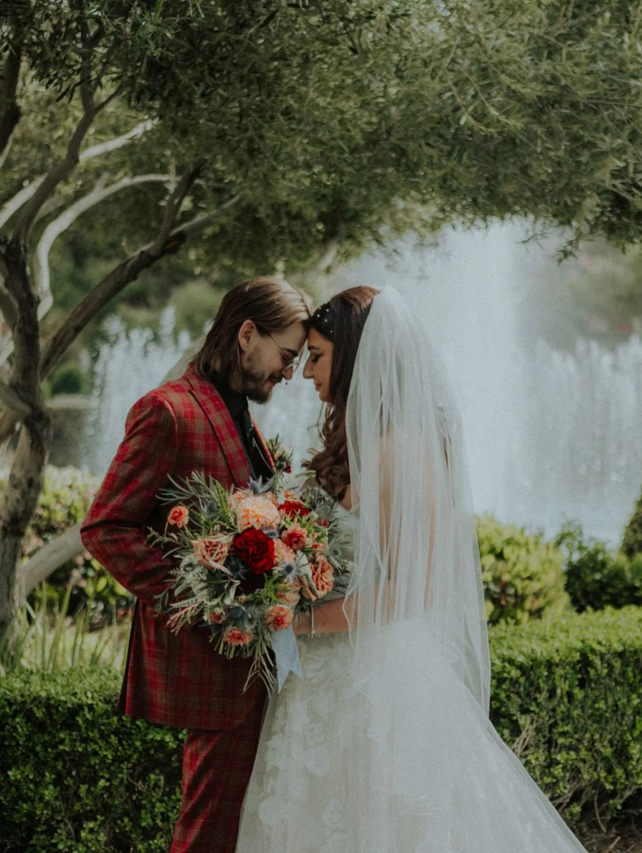 Bride and groom standing close together outdoors, with foreheads touching, holding a bouquet of flowers, under a tree with water in the background.