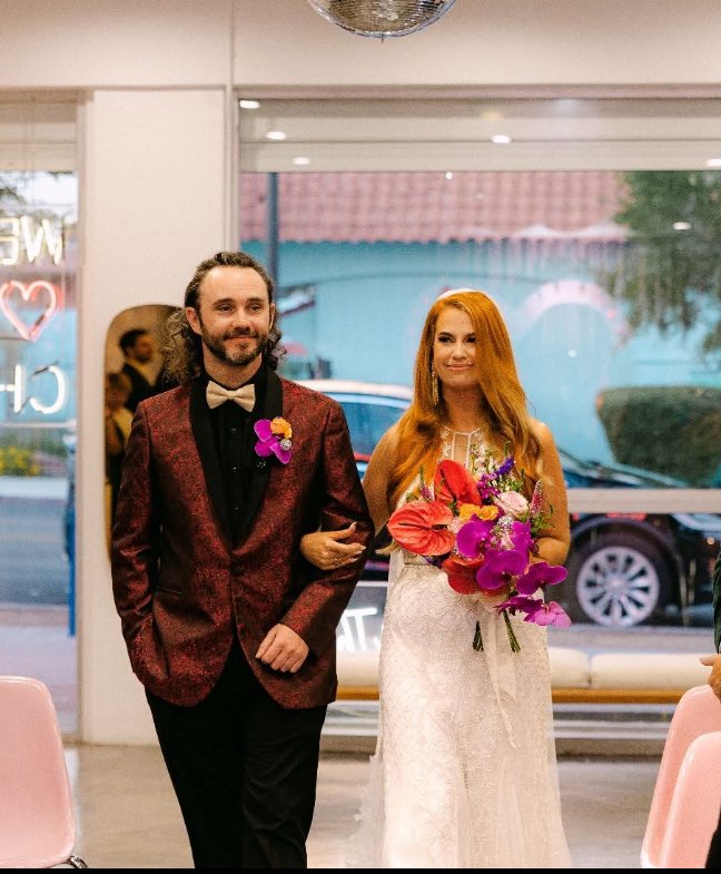 A bride with long red hair in a white wedding dress holding a colorful bouquet walking down the aisle, escorted by a man in a burgundy blazer with a white bowtie and flower pin, inside a well-lit venue with chairs and a window showing parked cars out