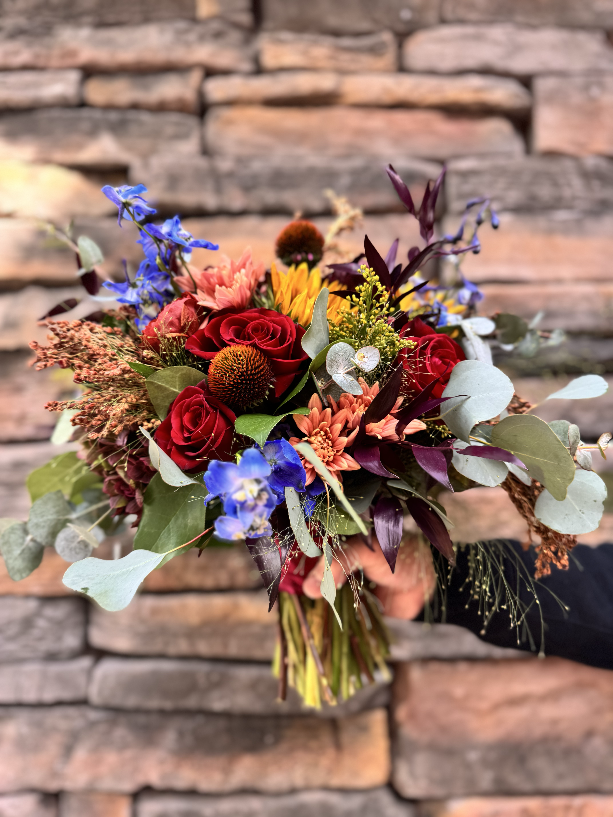 A colorful bouquet of flowers, including red roses, blue delphiniums, yellow sunflowers, orange chrysanthemums, and various green and purple leaves, held against a brick wall background.