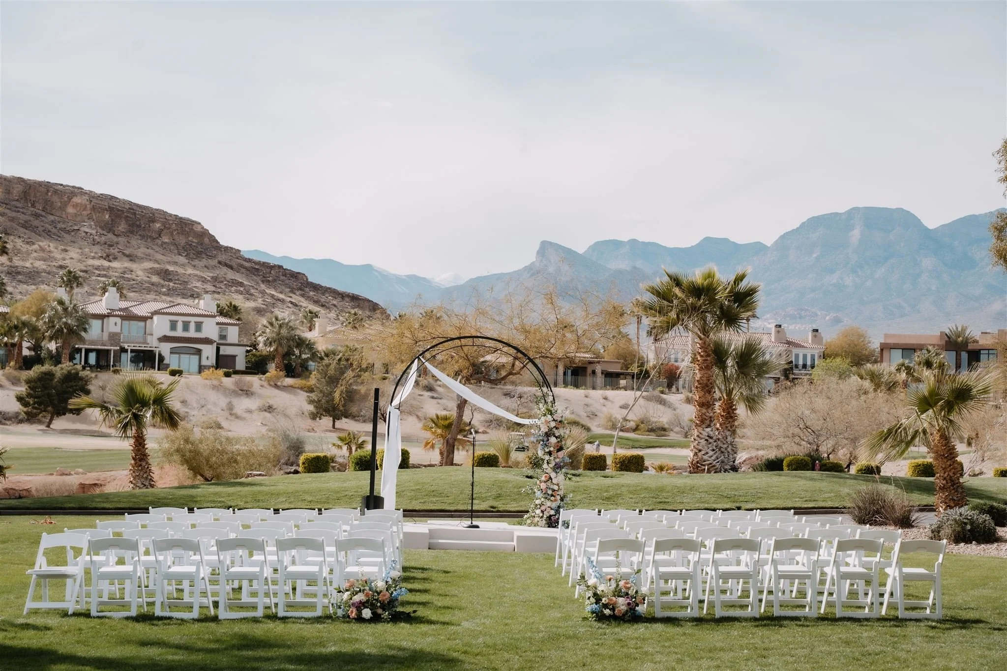 Outdoor wedding setup with white chairs arranged in rows on green grass, floral decorations at the aisle, a circular wedding arch with white drapery and flowers, palm trees, and mountainous landscape in the background.