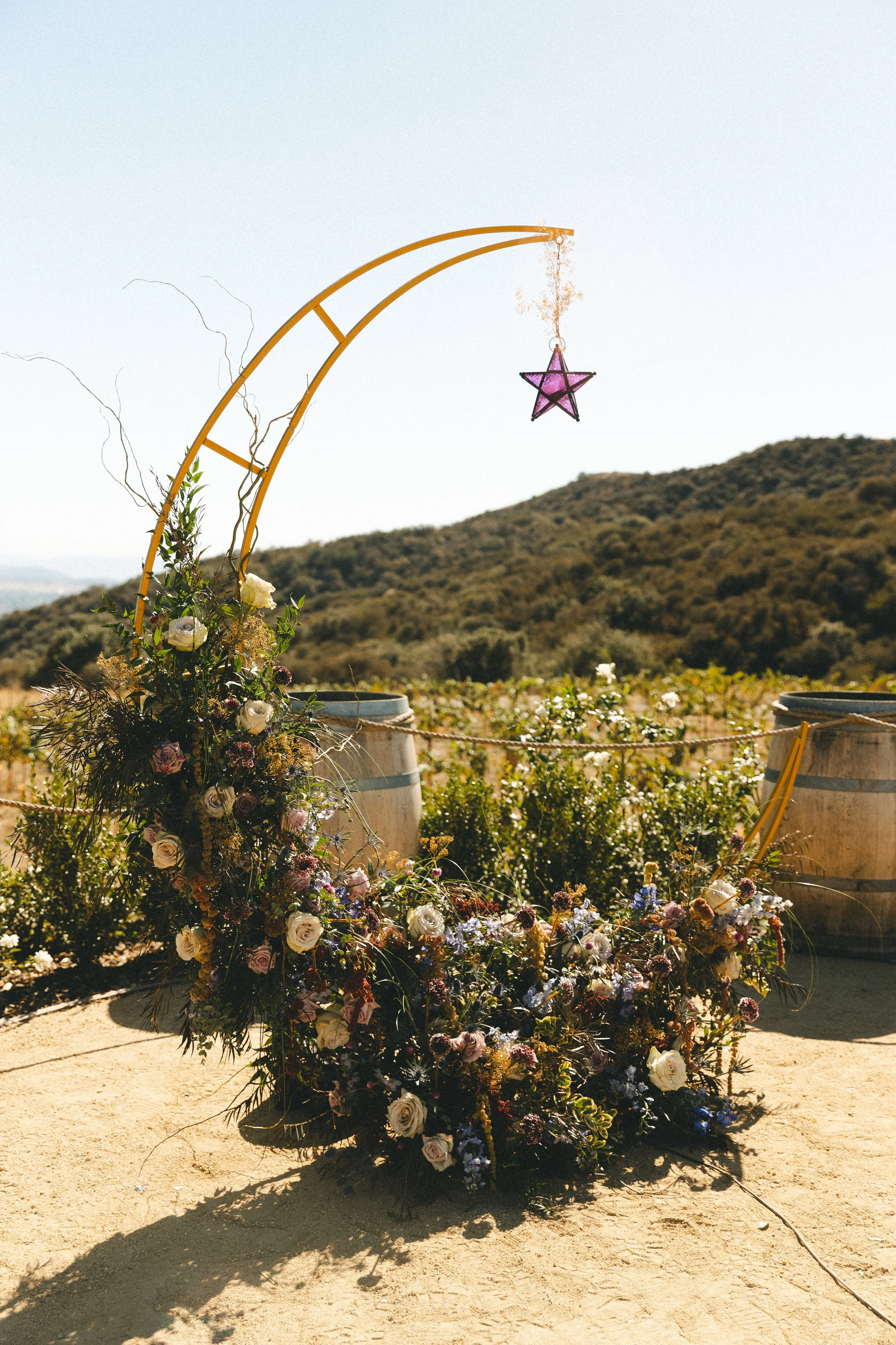 Decorative floral wedding arch on a dirt path with barrels and a scenic hillside in the background.