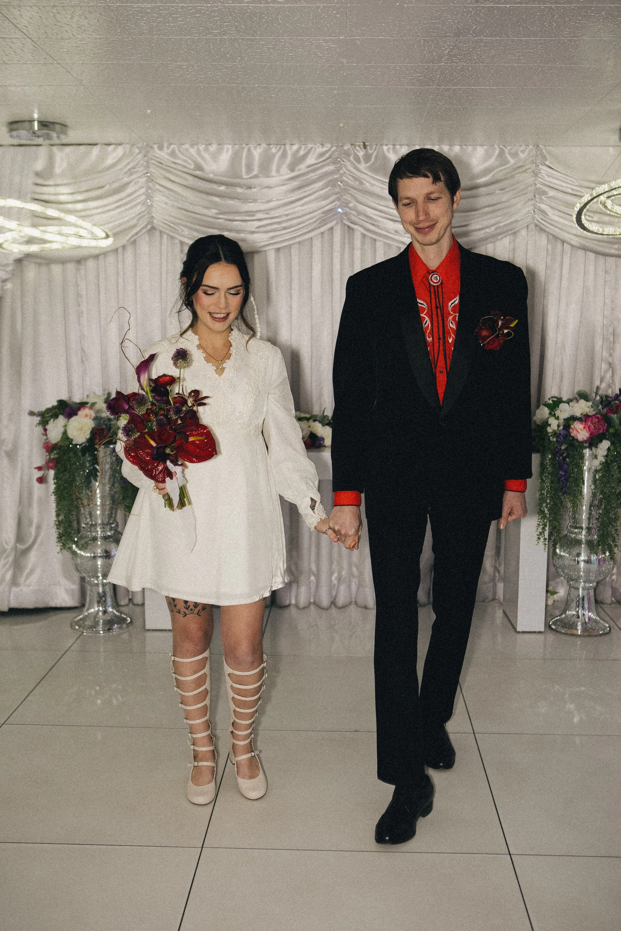 A bride and groom holding hands and walking at their wedding reception. The bride is wearing a white dress, beige heels, and holding a bouquet. The groom is wearing a black suit with a red shirt and decorative boutonniere. The background includes flo