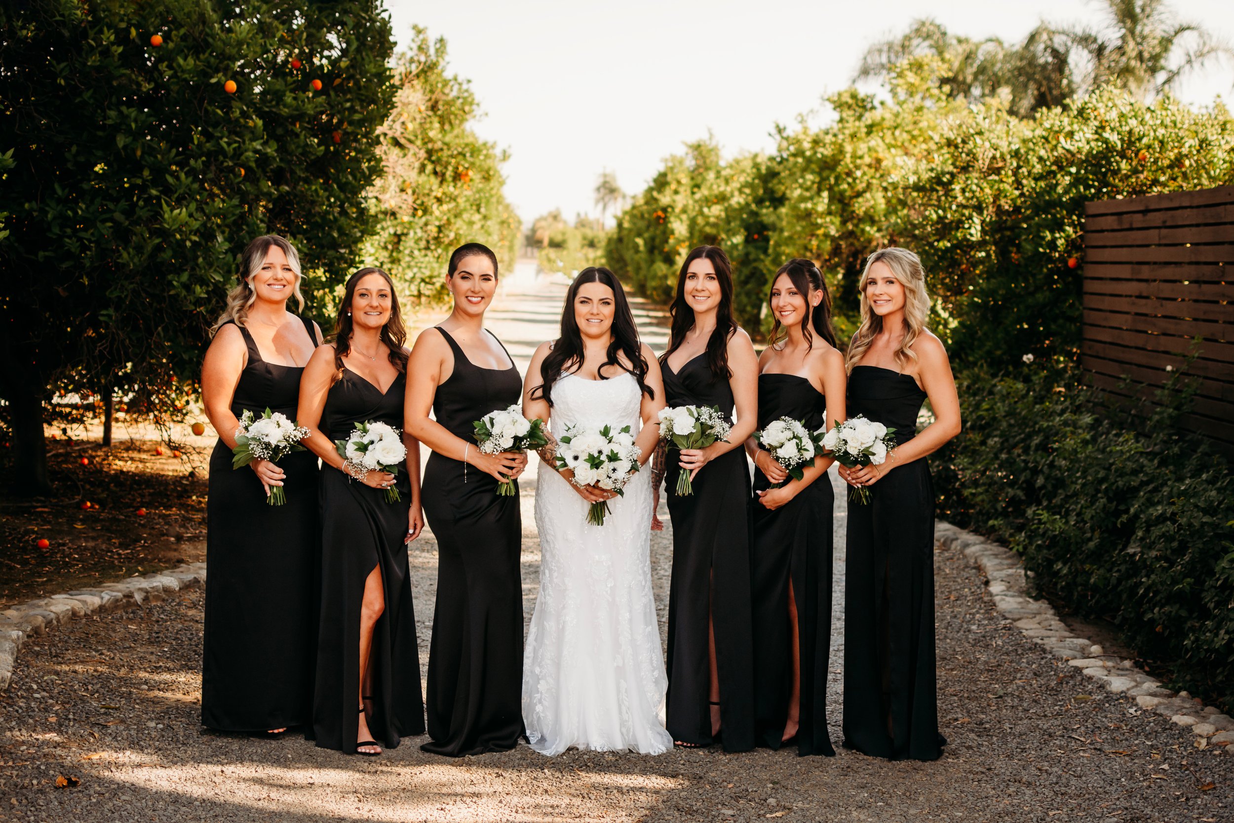 A bride in a white wedding dress standing with six bridesmaids in black dresses, holding white flower bouquets, outdoors on a sunny day with orange trees in the background.