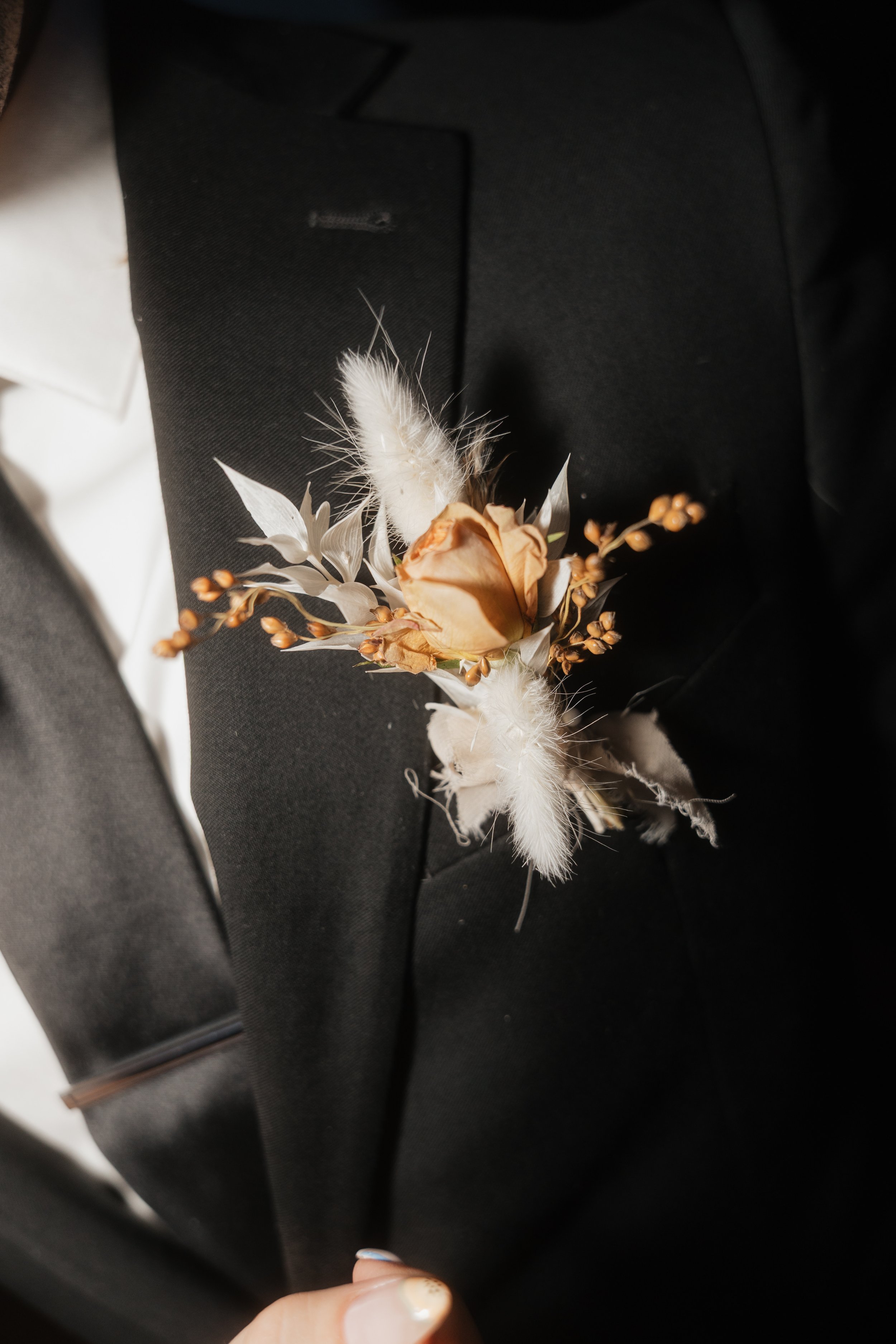 A boutonniere with dried flowers and feathers attached to a person's lapel on a black jacket.