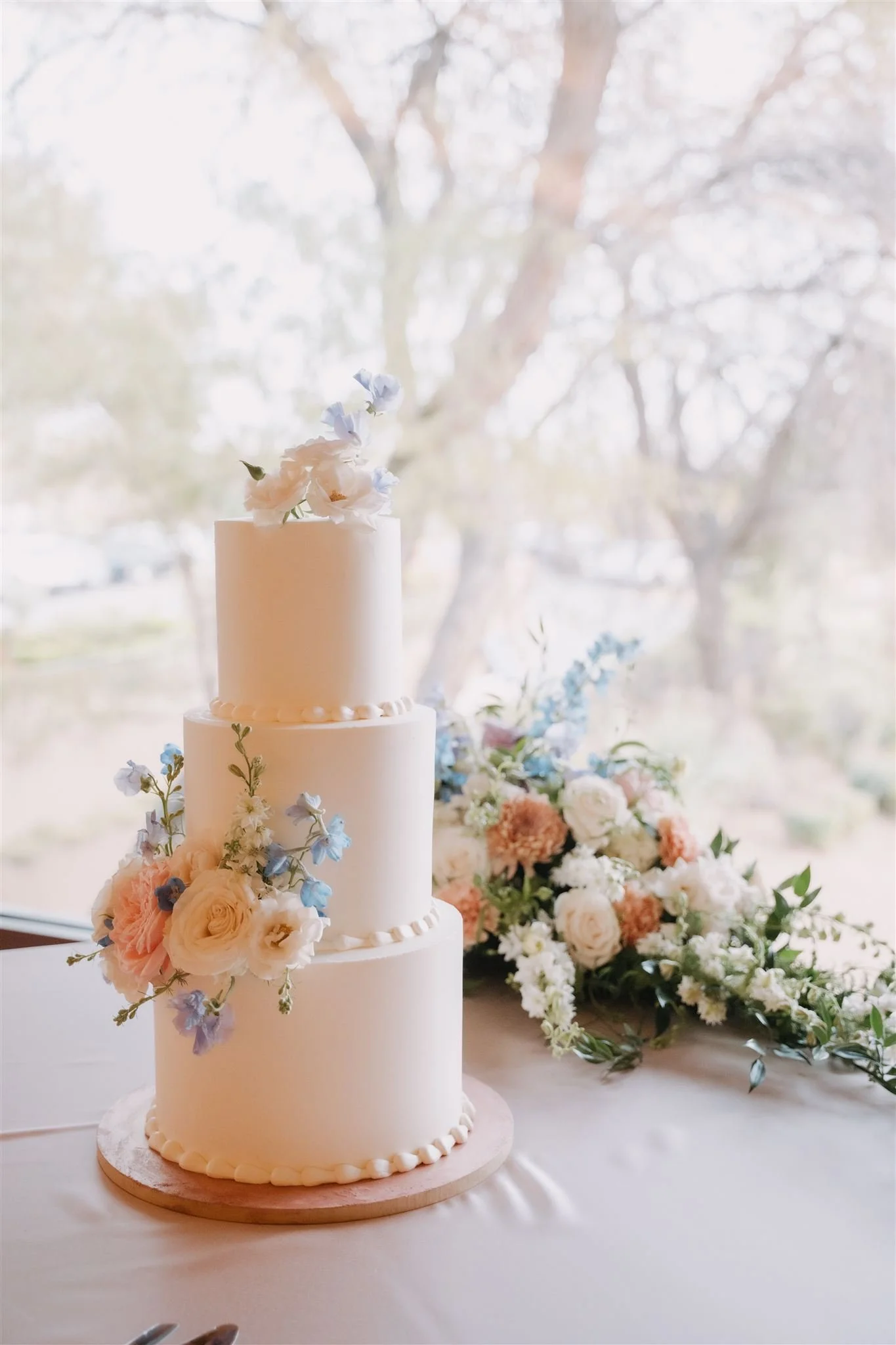 A three-tiered white wedding cake decorated with pink and white flowers, set on a wooden cake stand next to a bouquet of mixed flowers on a table with a white tablecloth. Sunlight filters through a window behind the cake.