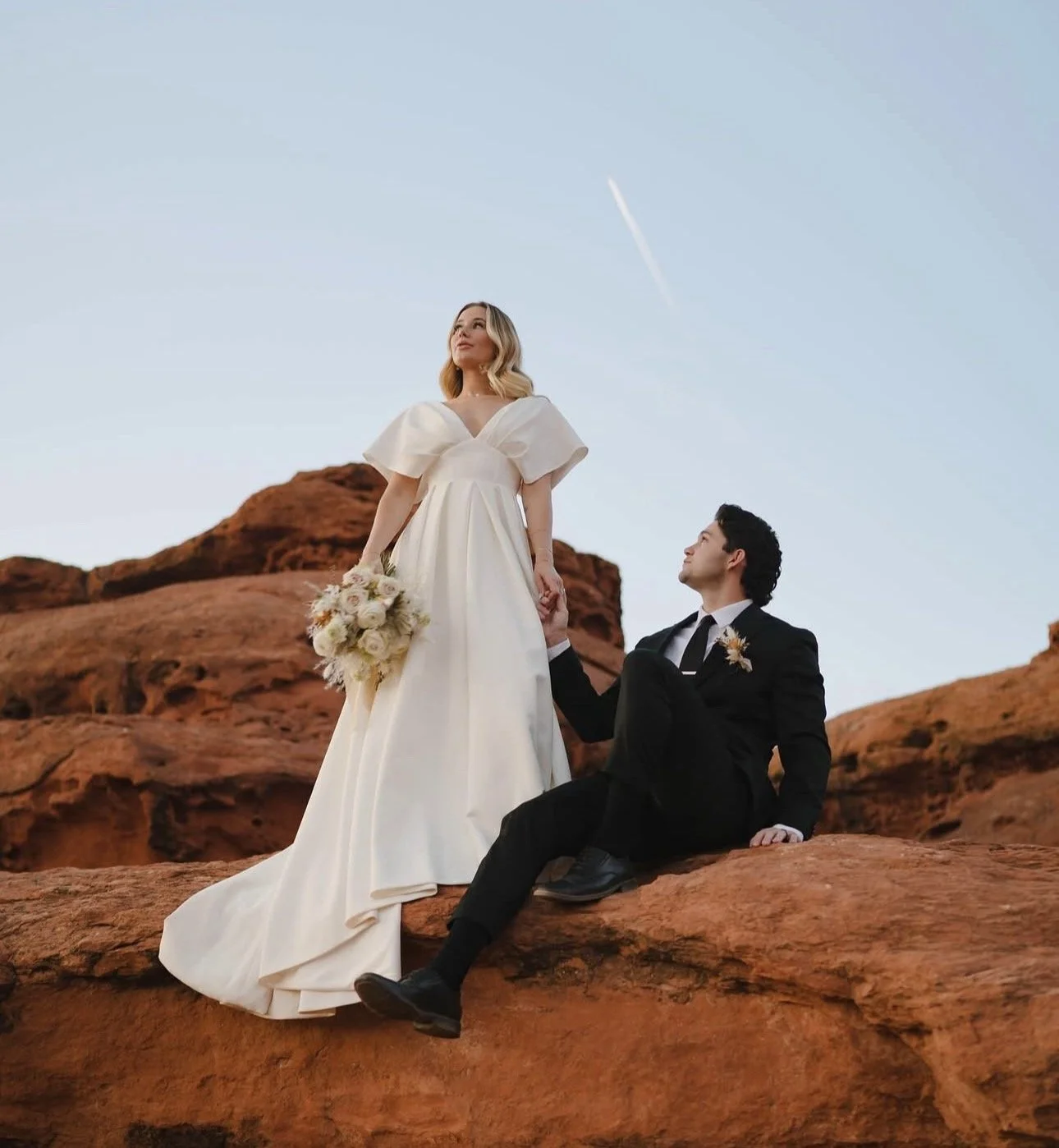 A bride and groom on a rocky desert landscape at sunset. The bride is standing in a white wedding gown holding a bouquet, and the groom is sitting on the rocks looking up at her.