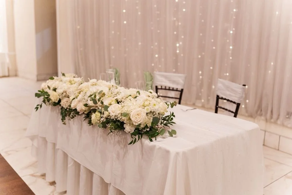 A wedding reception table decorated with white flowers and greenery, white tablecloth, and chairs with white covers, set against a backdrop of draped curtains with twinkling lights.