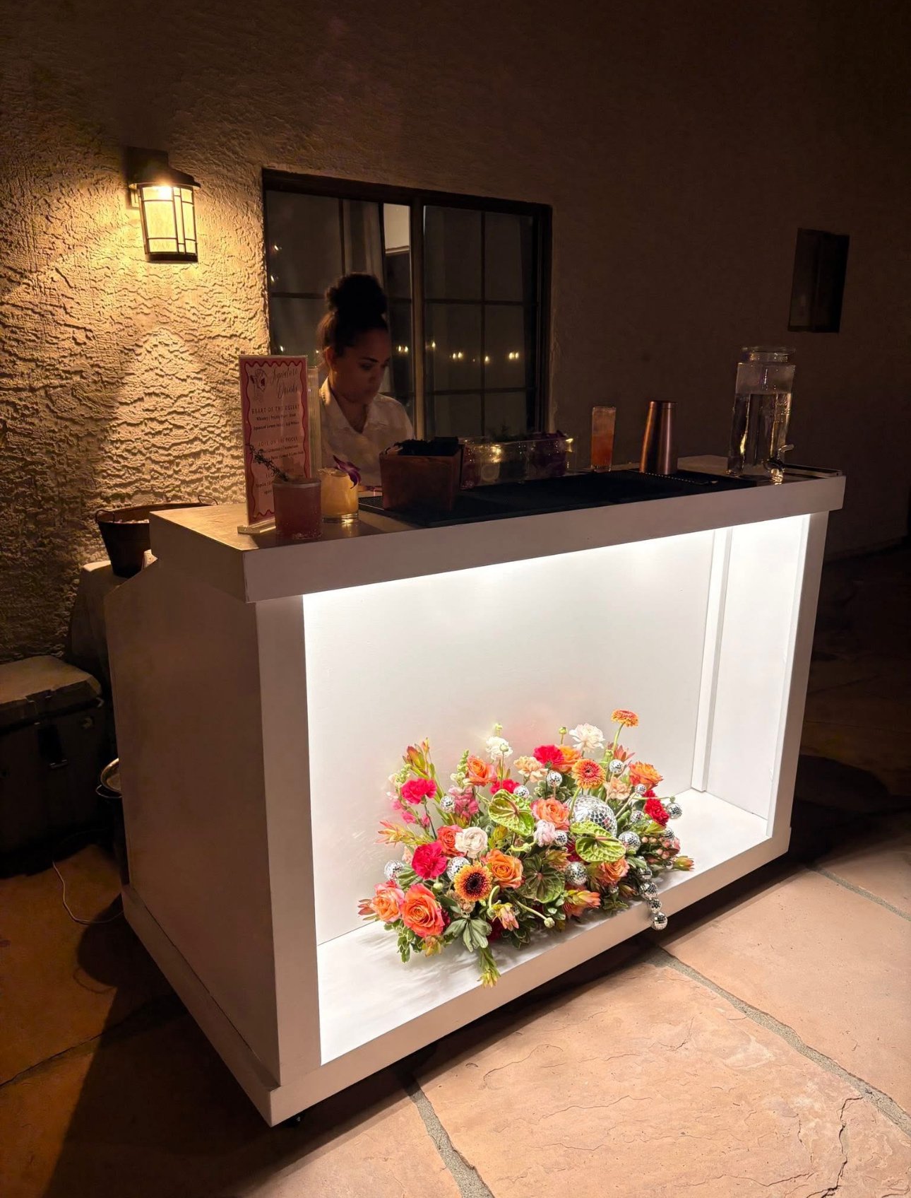 A white illuminated bar with a floral arrangement at the bottom, and a woman behind it, in a dimly lit setting.