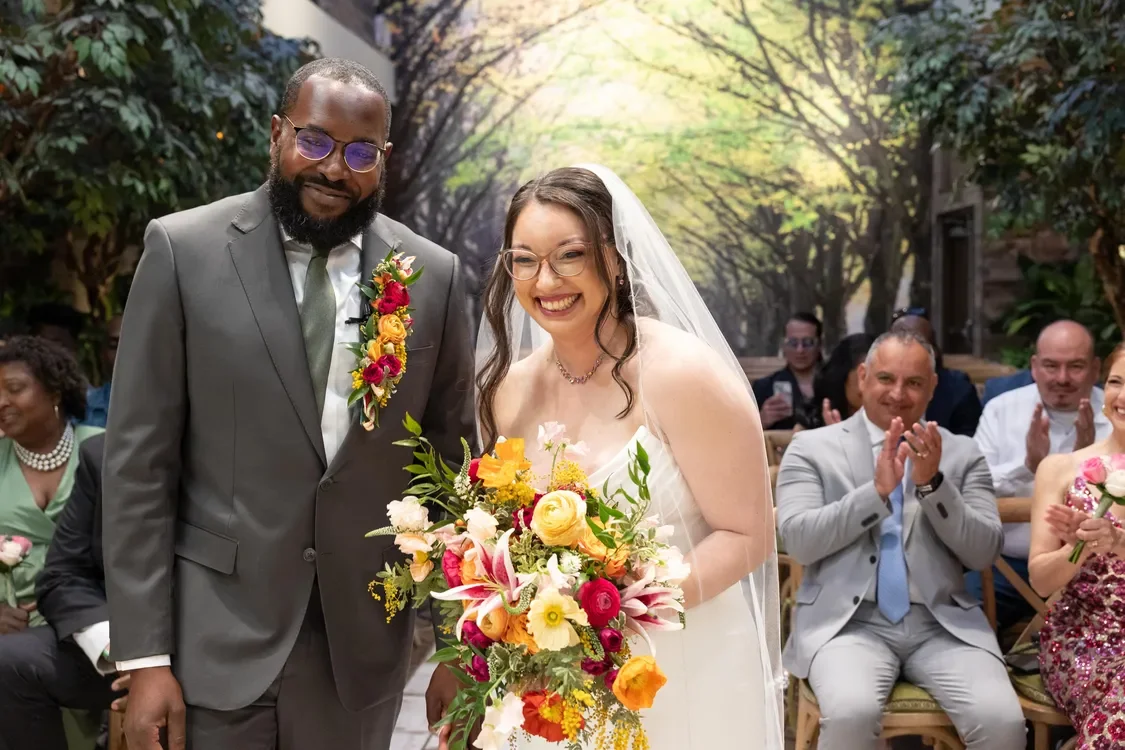 A bride and groom standing together at their wedding ceremony, smiling and holding a vibrant bouquet of flowers, with wedding guests seated and clapping in the background.
