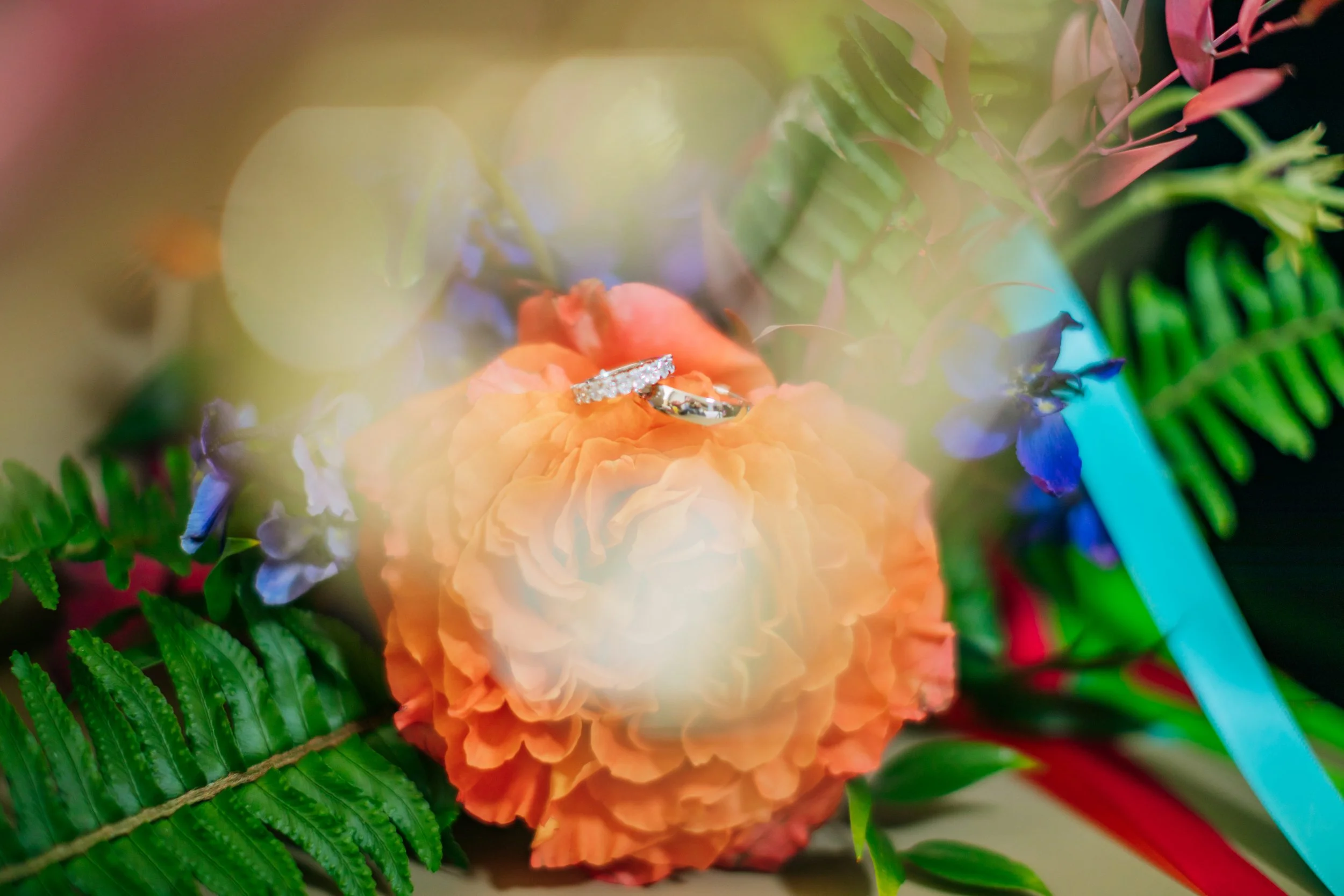 Diamond engagement ring placed on an orange flower surrounded by green leaves, purple flowers, and a blue ribbon.