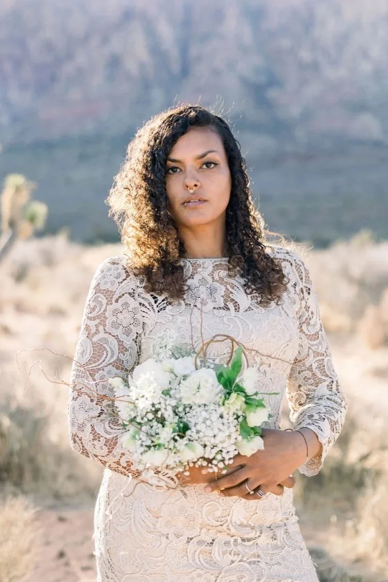 A woman with curly hair wearing a lace dress, holding a bouquet of white flowers, standing outdoors in a desert landscape with mountains in the background.