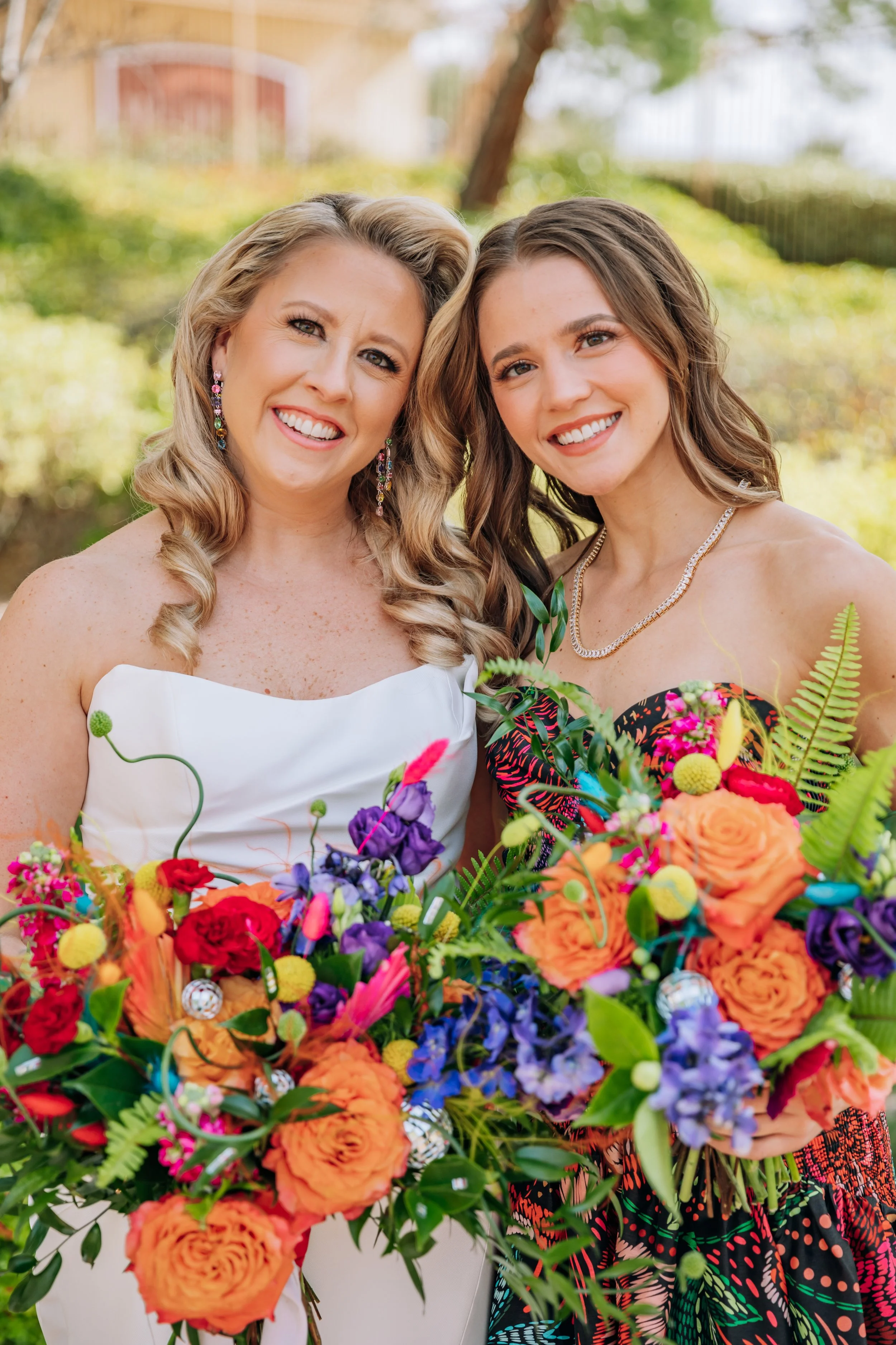 Two women smiling outdoors, holding colorful bouquets of flowers, dressed in festive clothing.