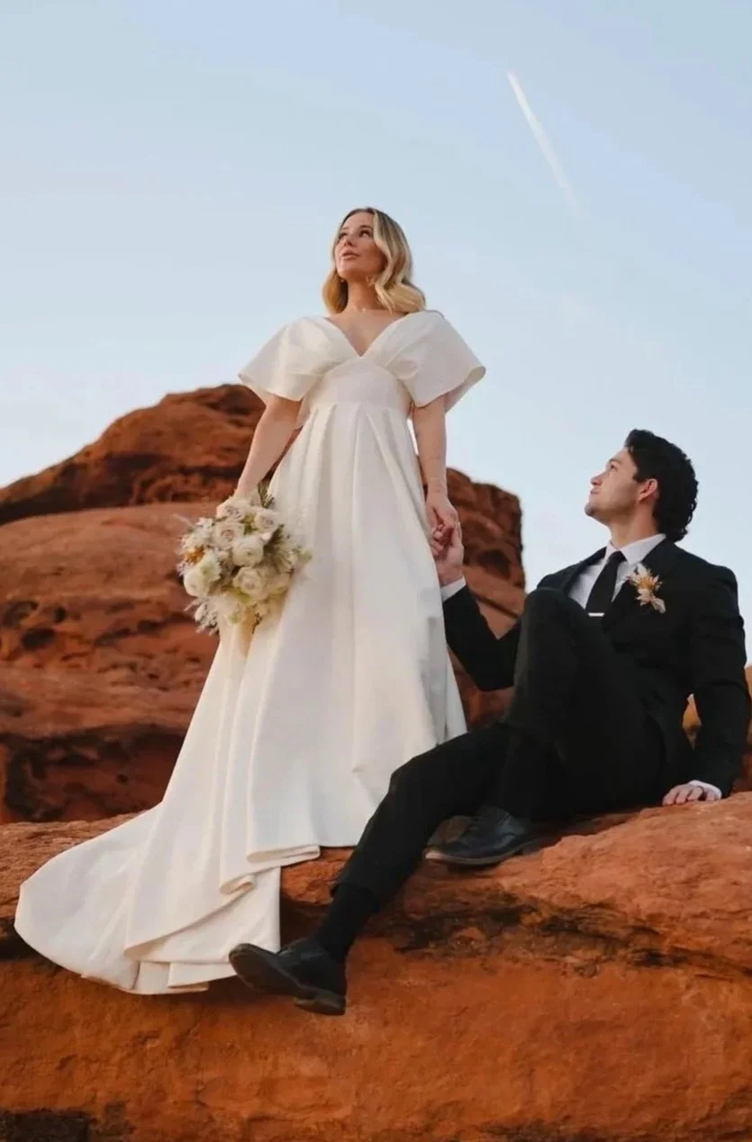 A bride in a white wedding dress holding a bouquet of flowers, standing on a rock formation, looking up, with a groom in a black suit sitting next to her, holding her hand, in a desert landscape during sunset or late afternoon.