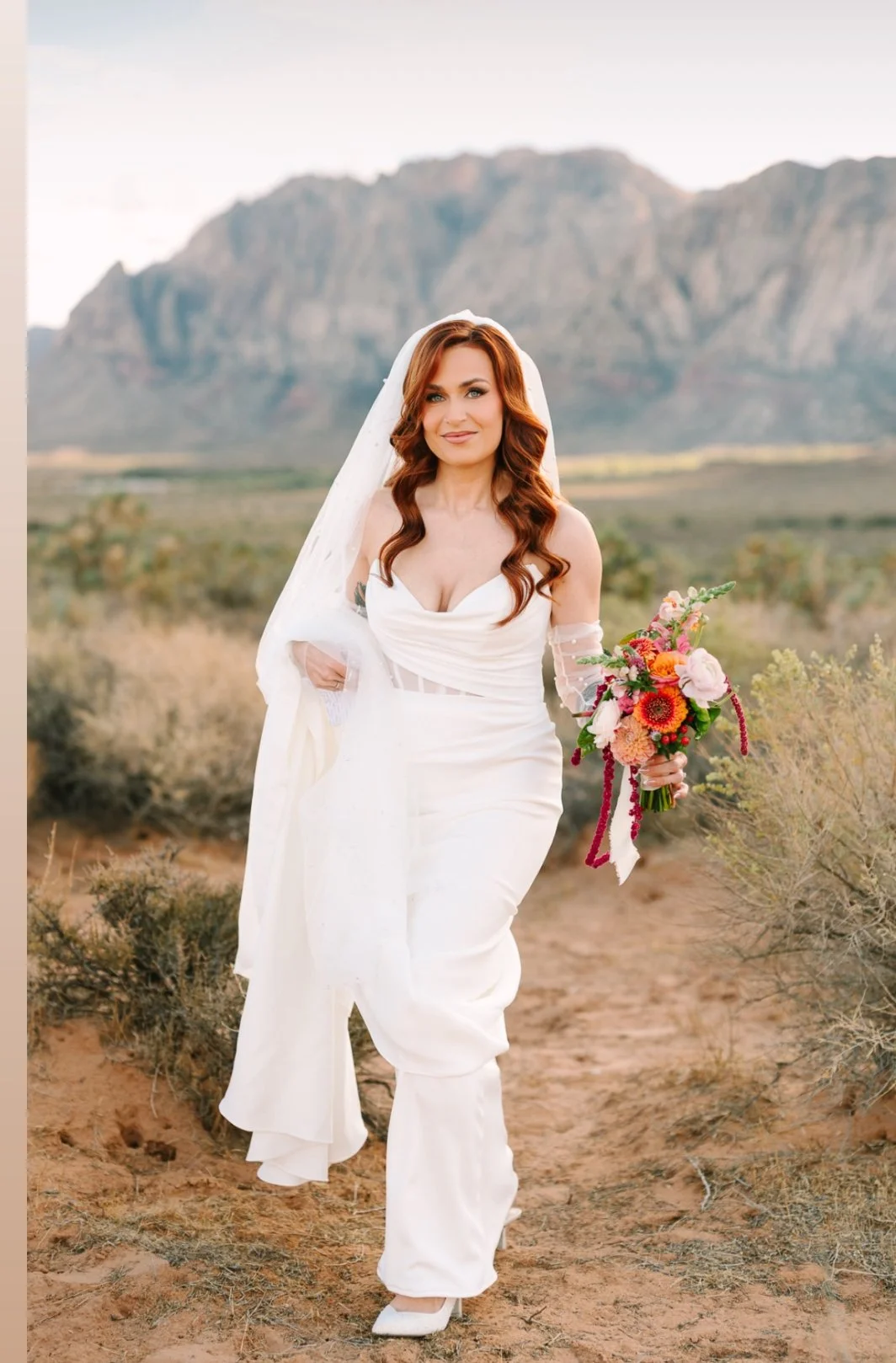 A woman with long, wavy red hair wearing a white wedding dress and veil, holding a colorful bouquet, standing outdoors in a desert landscape with mountains in the background.