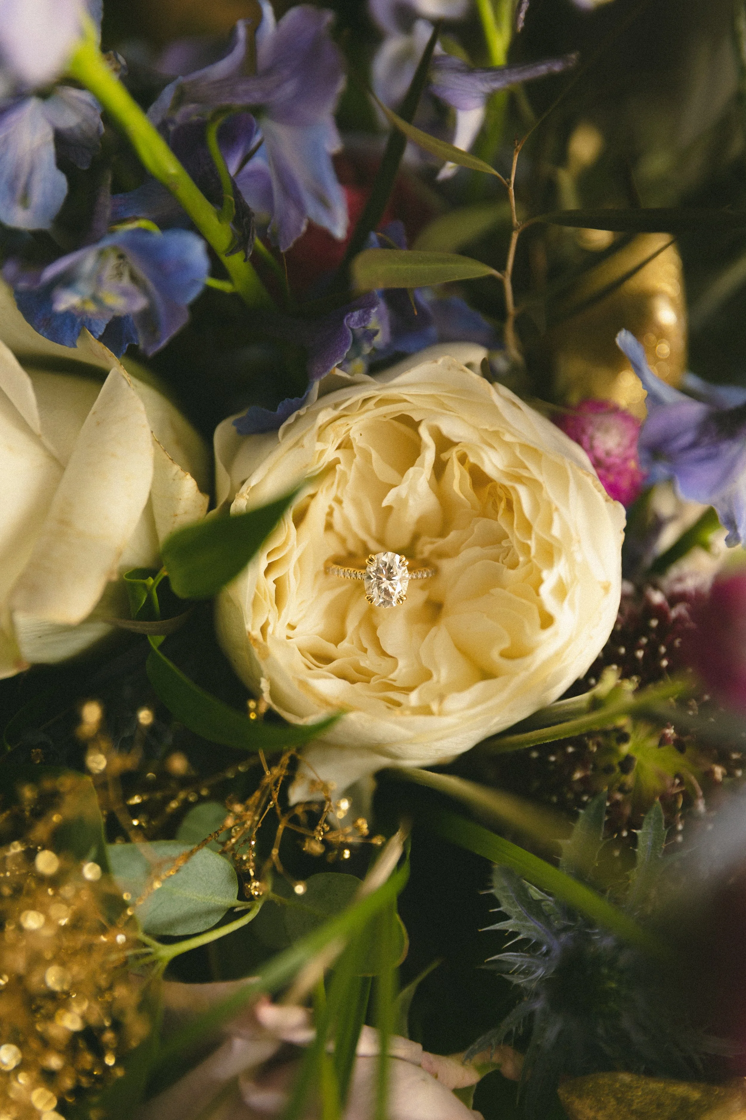 A diamond engagement ring resting inside a cream-colored flower surrounded by purple, pink, and green floral elements.