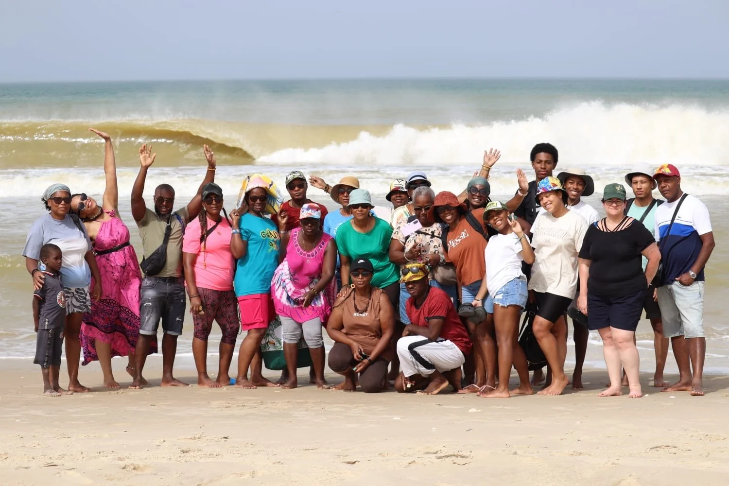 Groupe de personnes posant sur la plage avec l'océan en arrière-plan, certaines portant des chapeaux, des lunettes de soleil et des vêtements de plage.