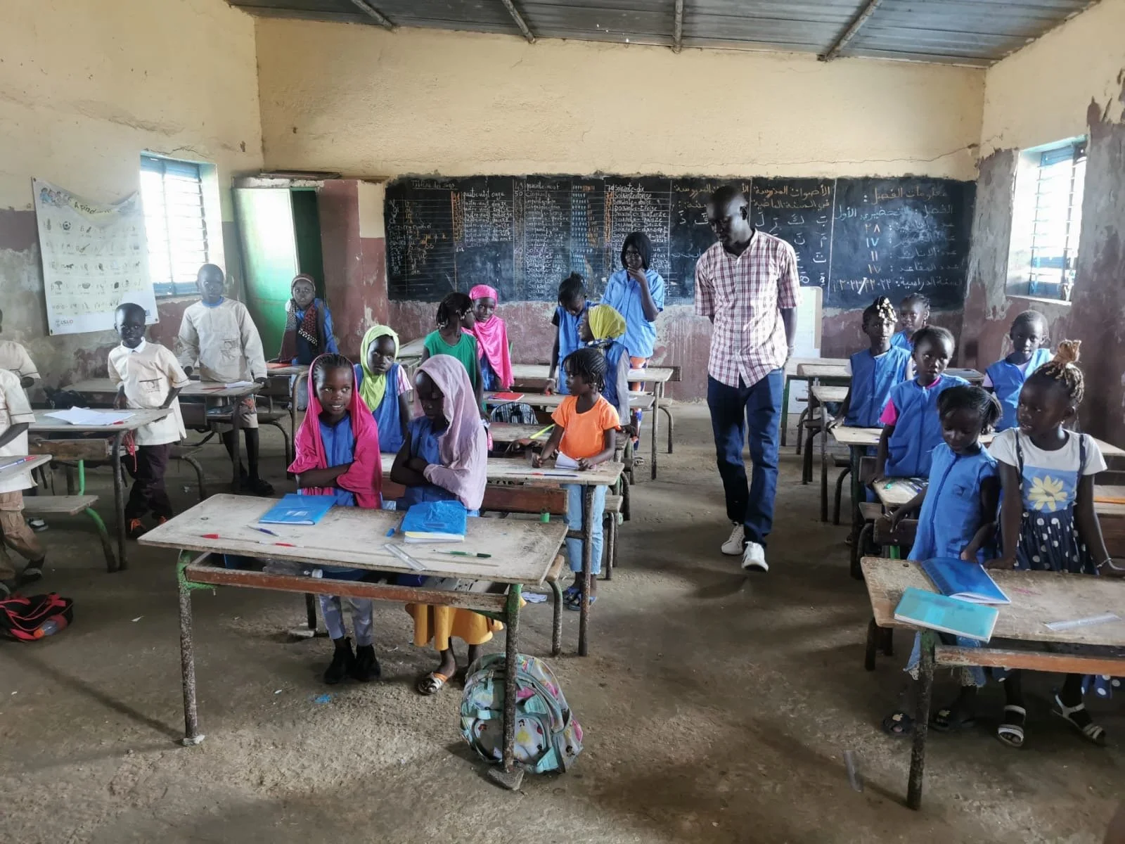 Une classe scolaire avec des élèves et un professeur, dans une salle en mauvais état avec des murs abîmés et deux fenêtres. Les élèves portent des uniformes colorés et des livres sont sur les desks.