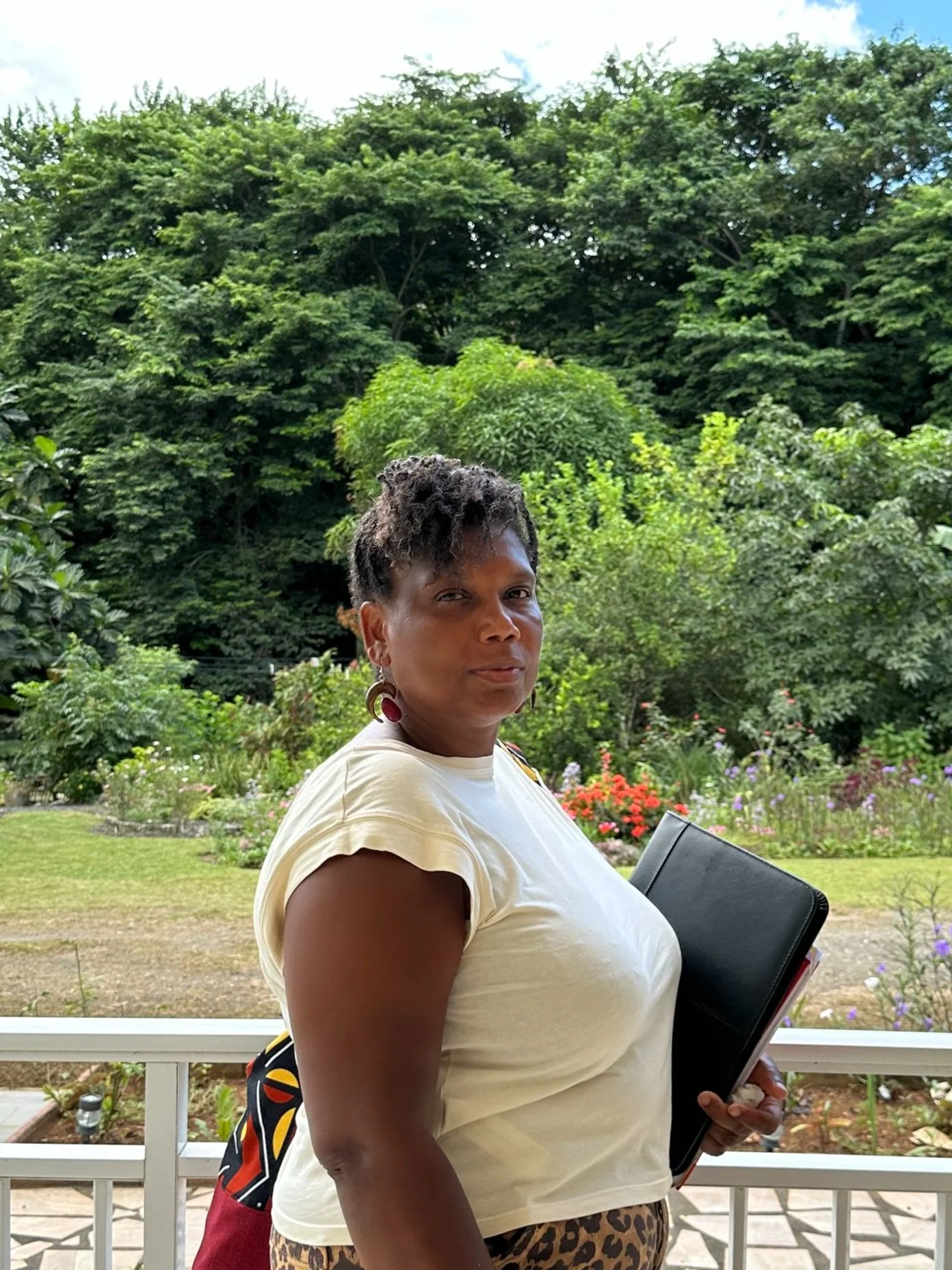 Une femme avec des cheveux courts et bouclés, portant un t-shirt blanc, des boucles d'oreilles rouges et un pantalon à motif léopard, tenant un classeur noir, se tenant sur une terrasse avec un jardin verdoyant en arrière-plan.