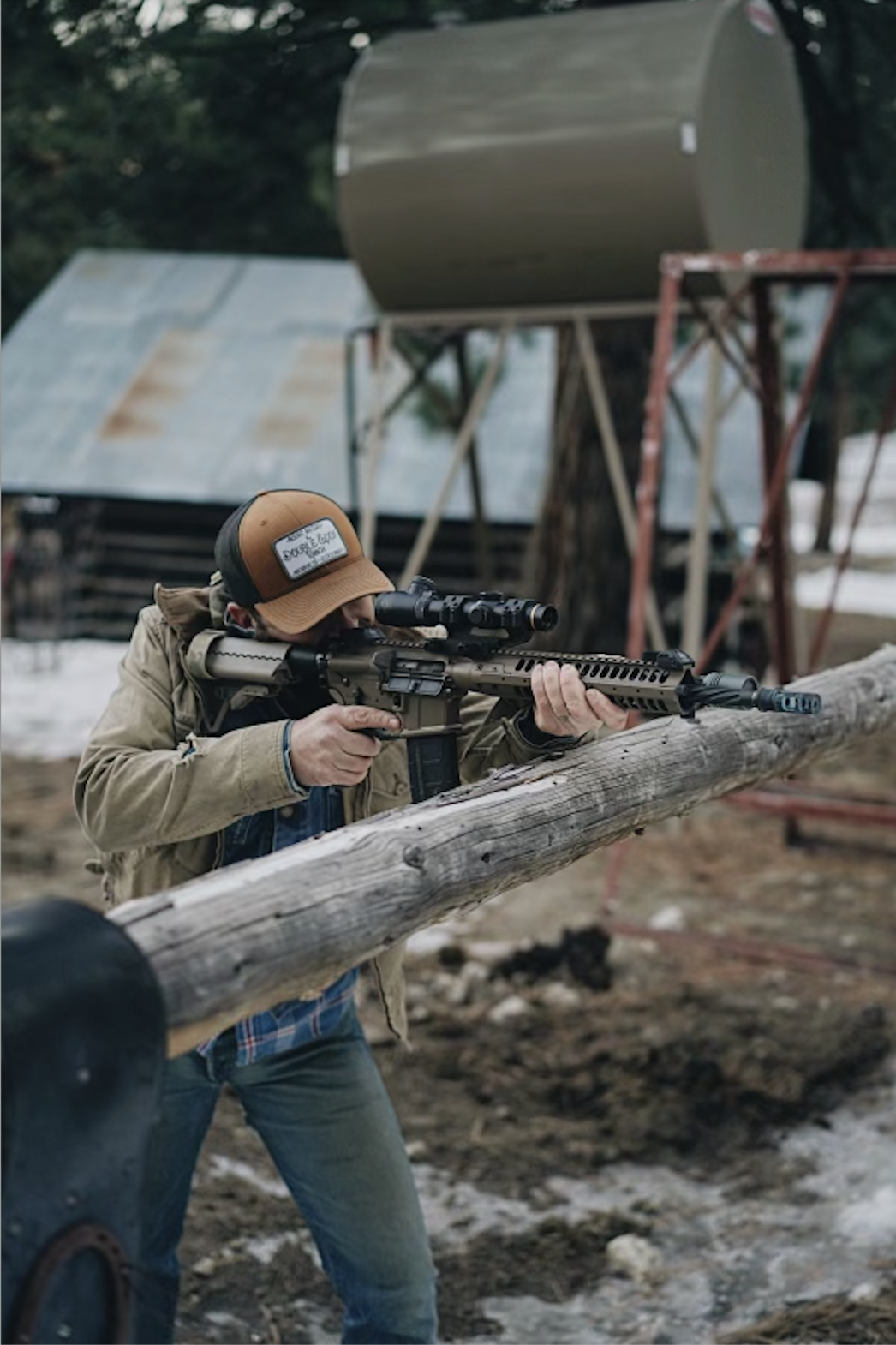 A man aiming a scoped rifle while kneeling behind a log outdoors with a rustic shed and trees in the background.