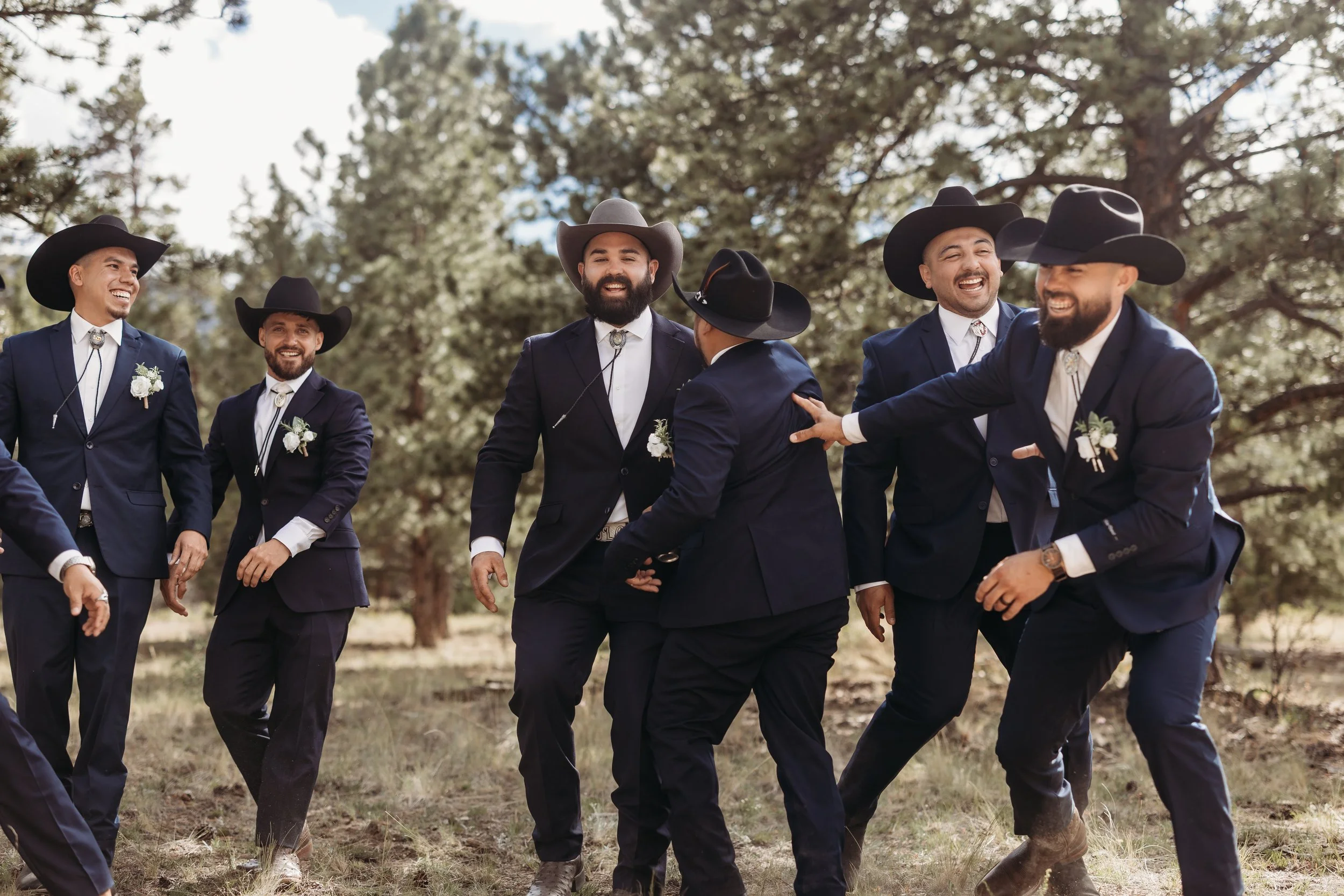 Group of men dressed as cowboys enjoying time outdoors, wearing dark suits, cowboy hats, and boutonnieres, with trees in the background.