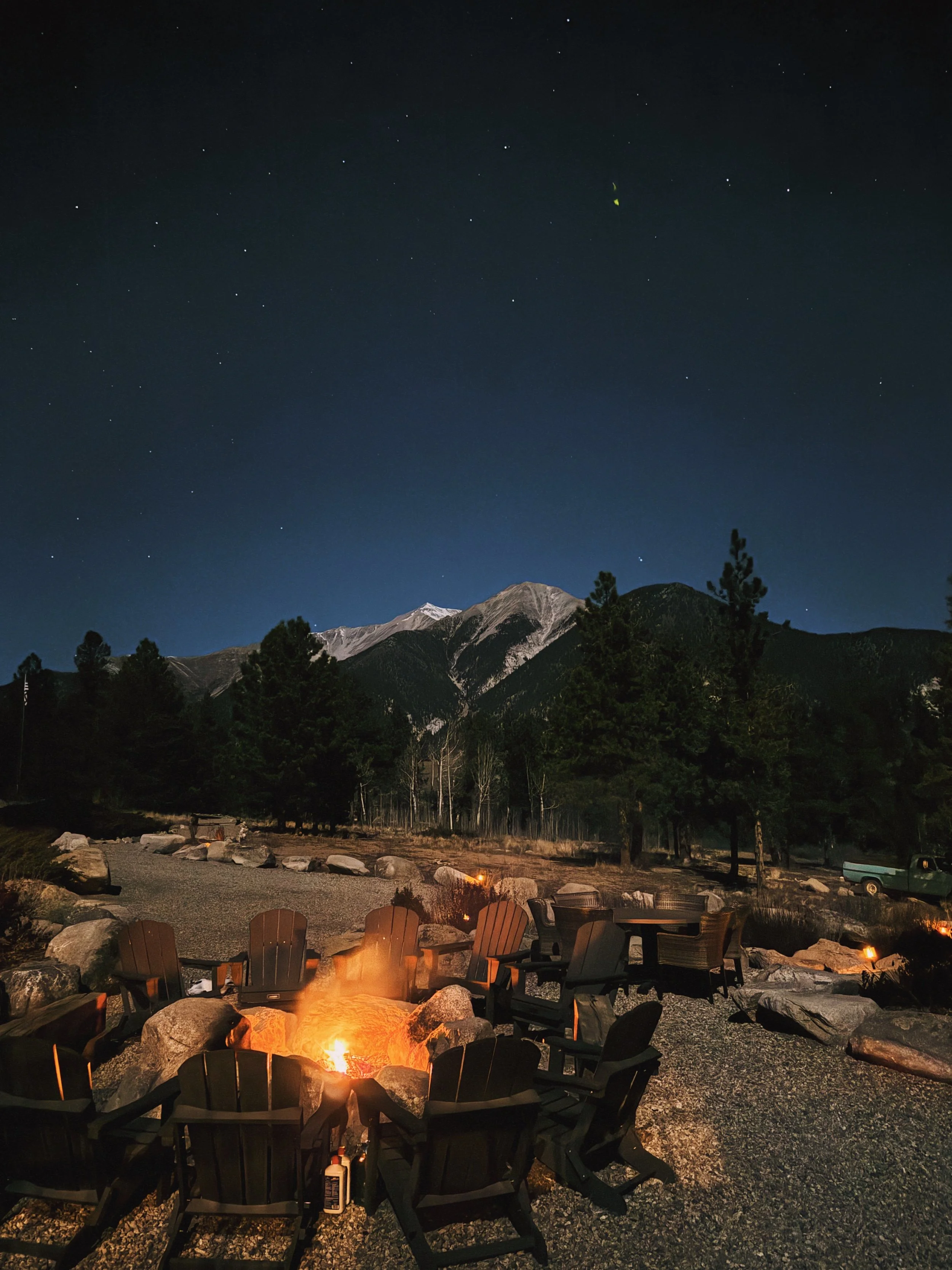Nighttime scene with a campfire surrounded by chairs, rocky terrain, and pine trees, with mountains and a starry sky in the background.