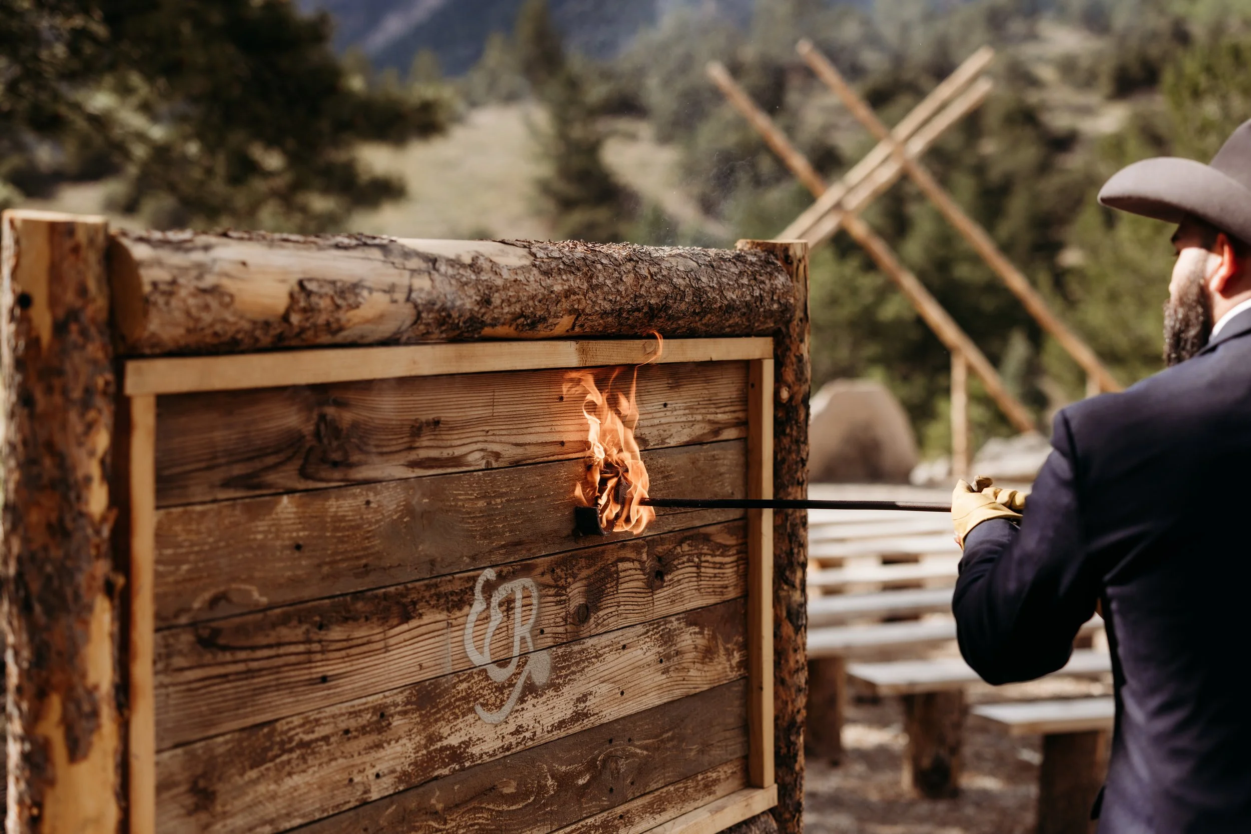 A man in a suit and cowboy hat igniting a wooden feature with a torch at an outdoor event, with mountains and trees in the background.