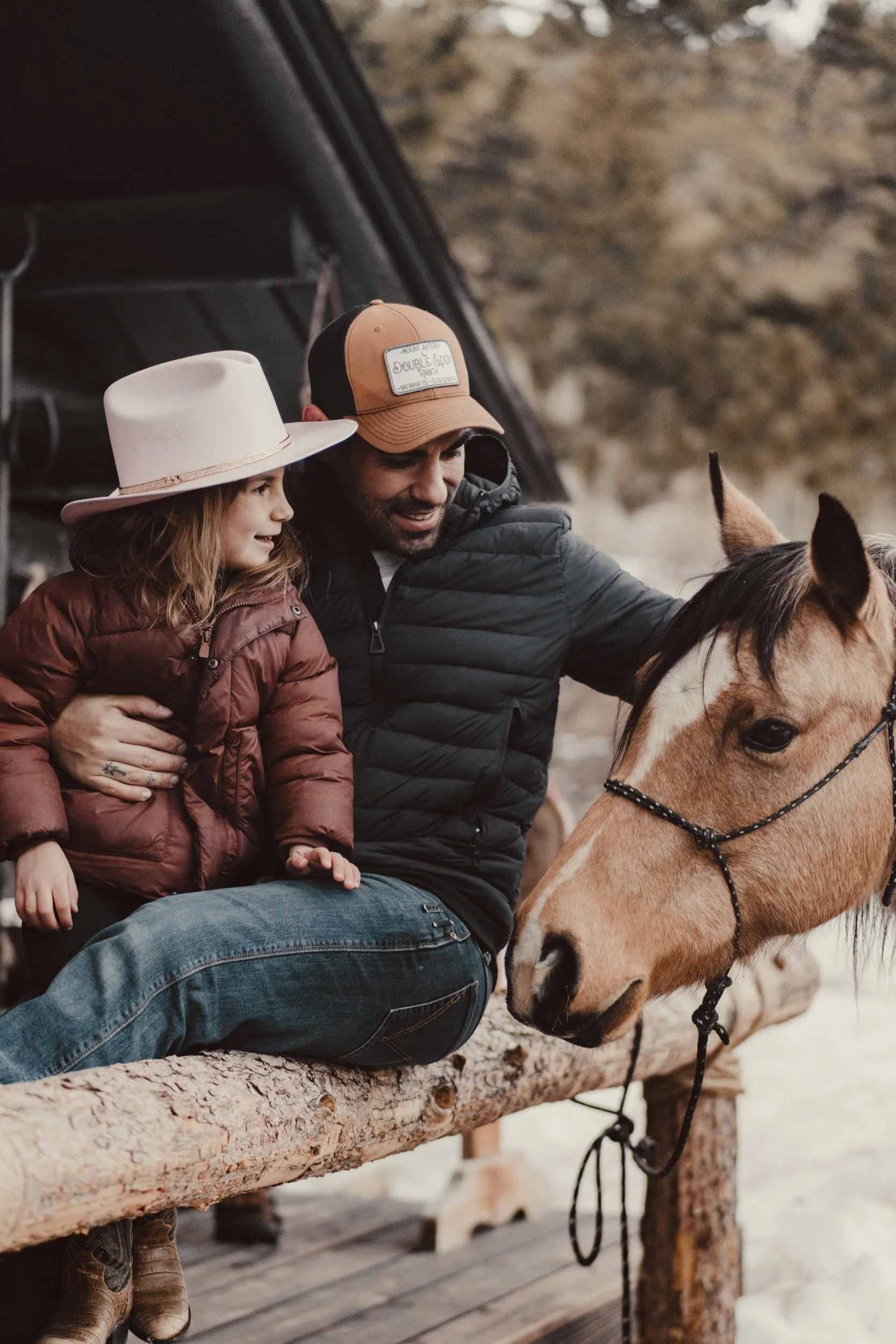 A man and a young girl sitting on a wooden railing outside, with a horse nearby. The girl is wearing a wide-brimmed hat and a brown puffer jacket, while the man is wearing a brown and black cap and a black jacket.