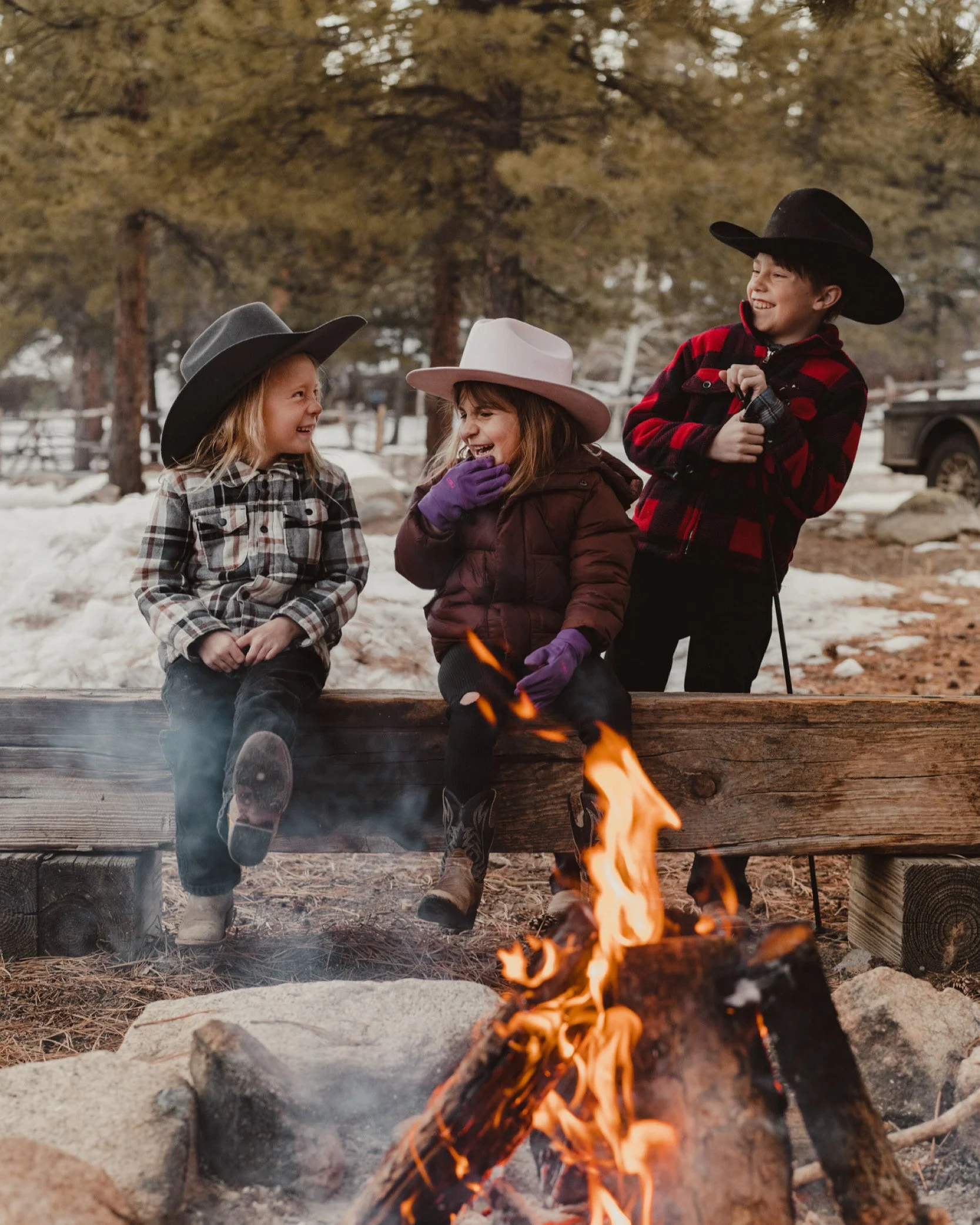 Three children wearing cowboy hats and winter clothes sitting and standing near a campfire in a snowy outdoor setting, smiling and laughing.