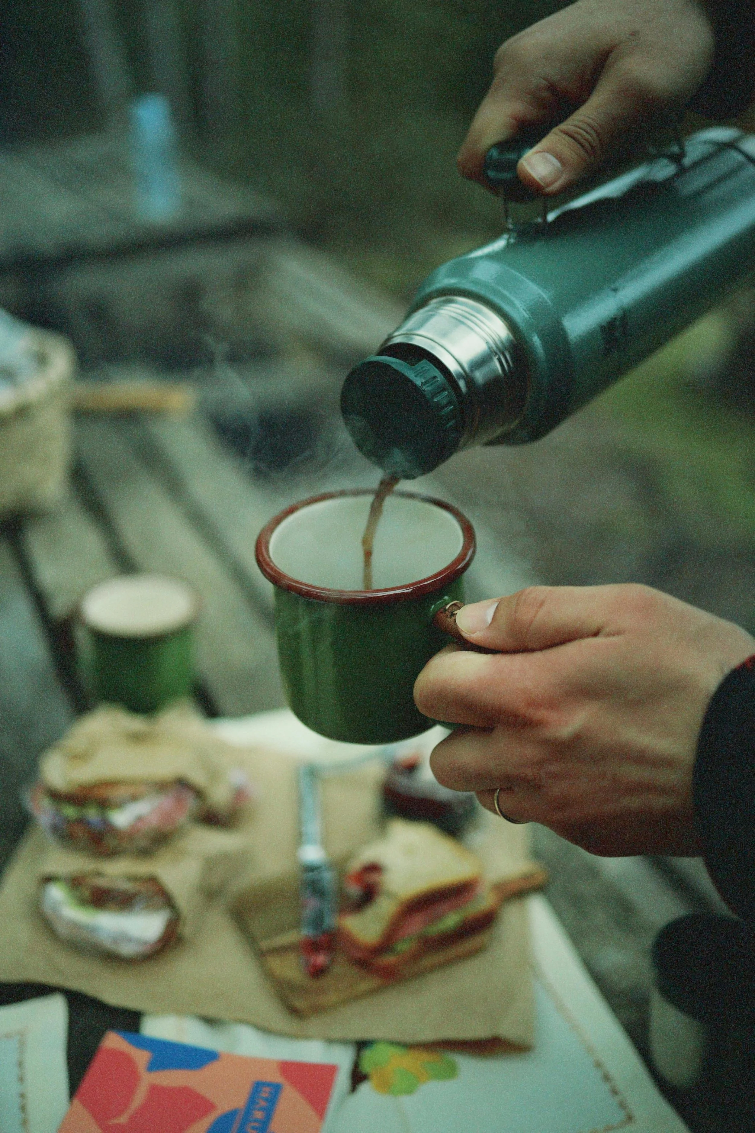 Person pouring hot coffee from a thermos into a green mug at an outdoor picnic table with sandwiches and snacks.