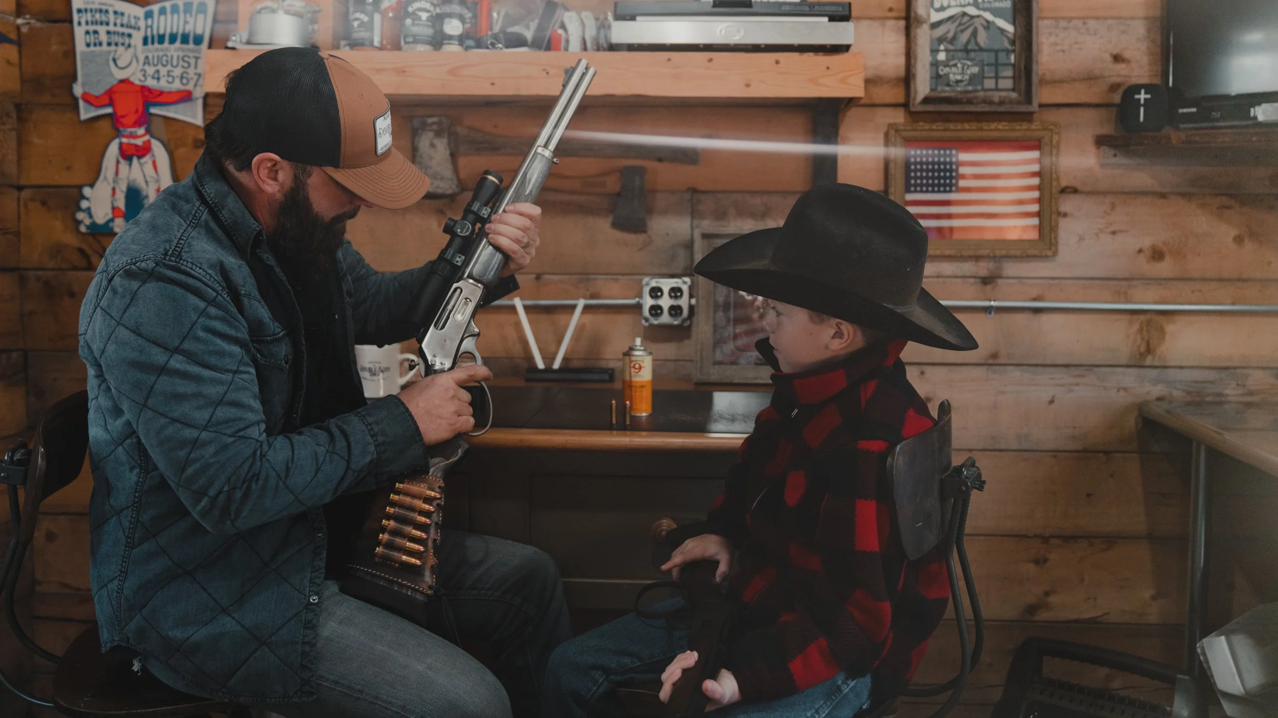 A man with a beard and glasses, wearing a denim jacket and a brown cap, is aiming a rifle at a young boy sitting on a chair. The boy is wearing a black cowboy hat and a red and black checkered jacket. The setting is a rustic wooden room with American-themed decorations on the wall.