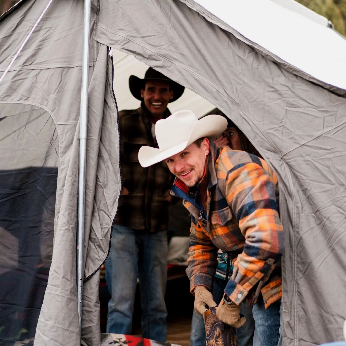 Three people inside a camping tent, two men wearing cowboy hats and a woman partially visible, smiling and looking into the camera.
