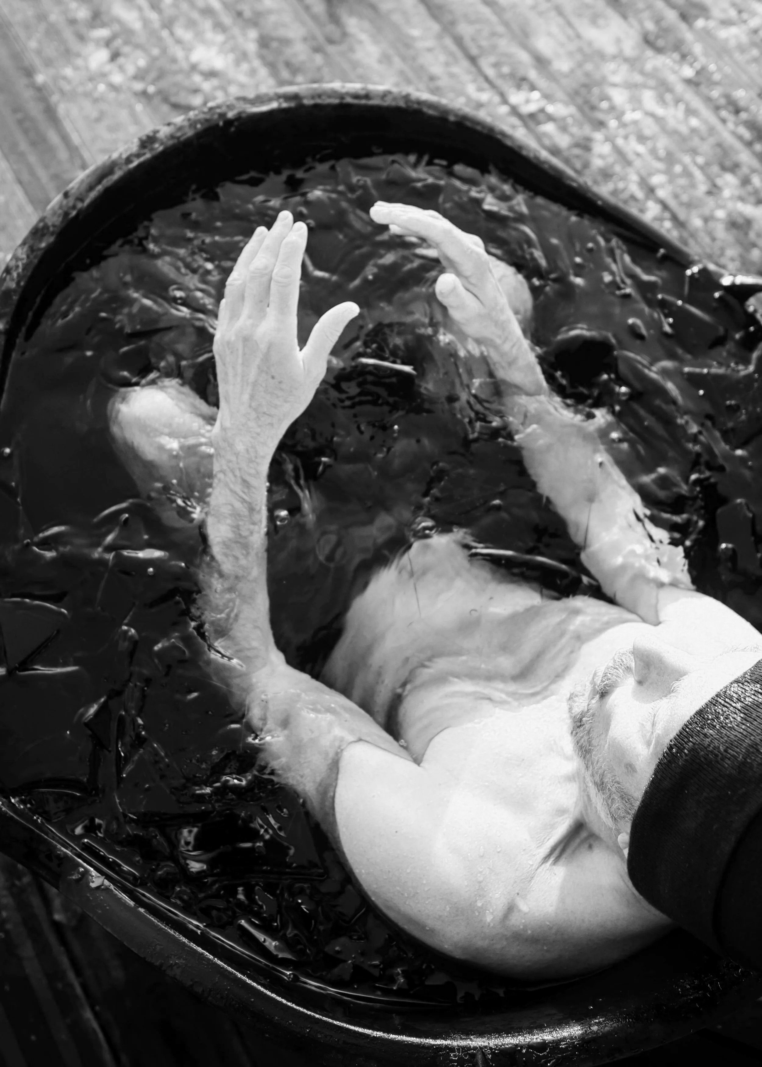 A person with a beard lying in a tub of water, with their hands raised near their face, and a dark cloth covering their eyes.