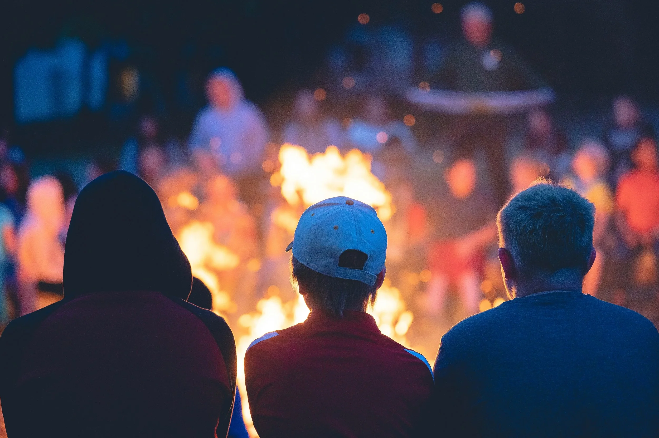 Three people sitting by a campfire at night, with a blurry background of other people and a boat, some with a person playing a guitar.