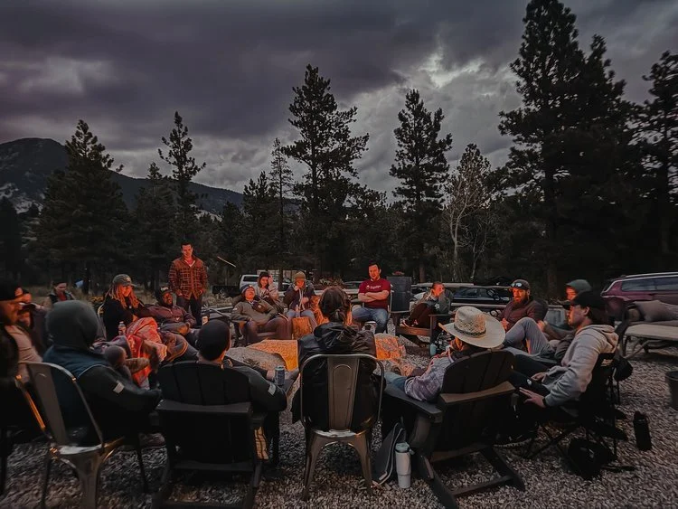A group of people gathered outdoors around a circle of chairs and blankets, possibly for a campfire or storytelling in a forested area with mountains and cloudy sky in the background.