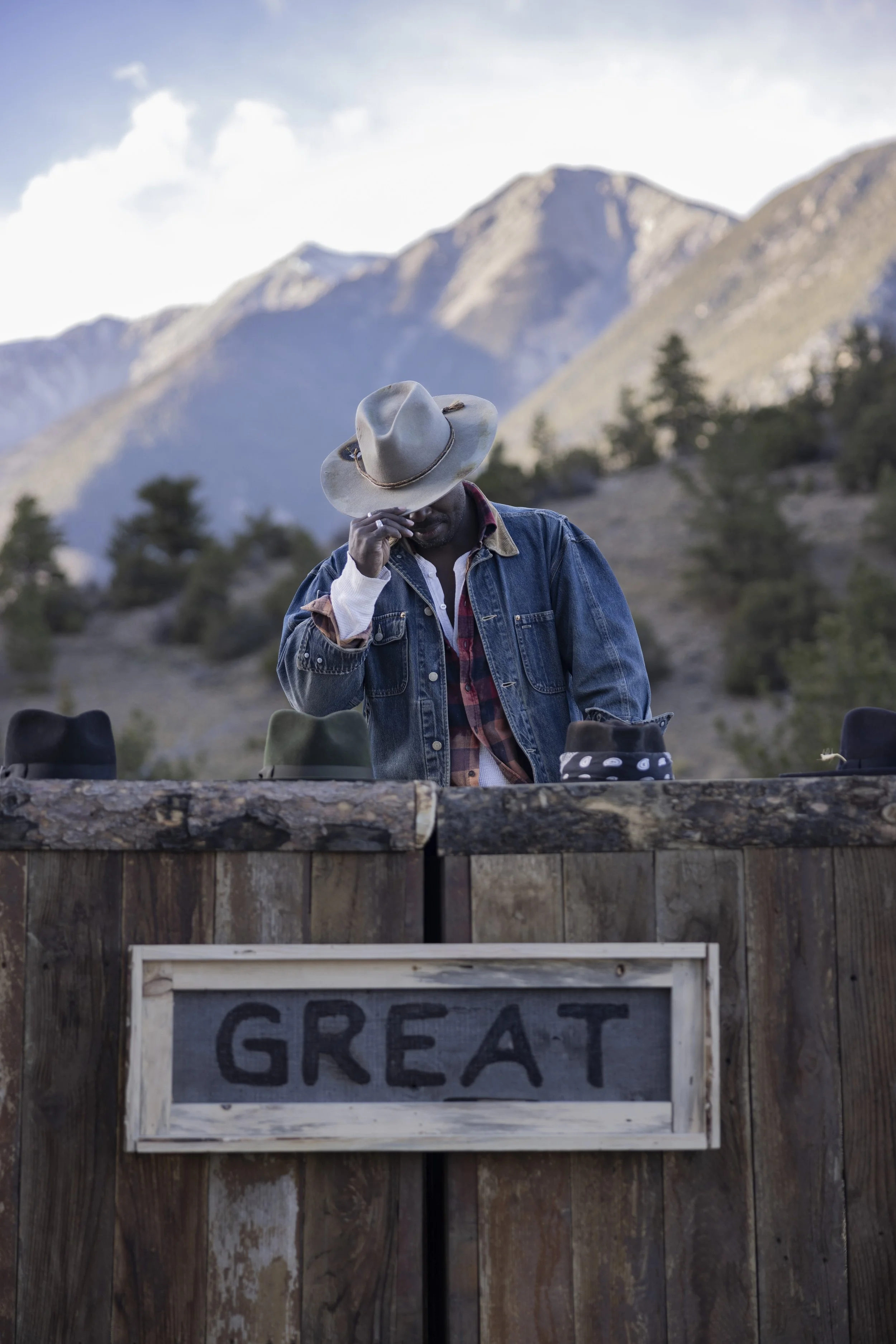 Man wearing a cowboy hat and denim jacket standing behind a wooden table outdoors with mountainous landscape in the background, with a sign that reads 'GREAT' in front of the table.