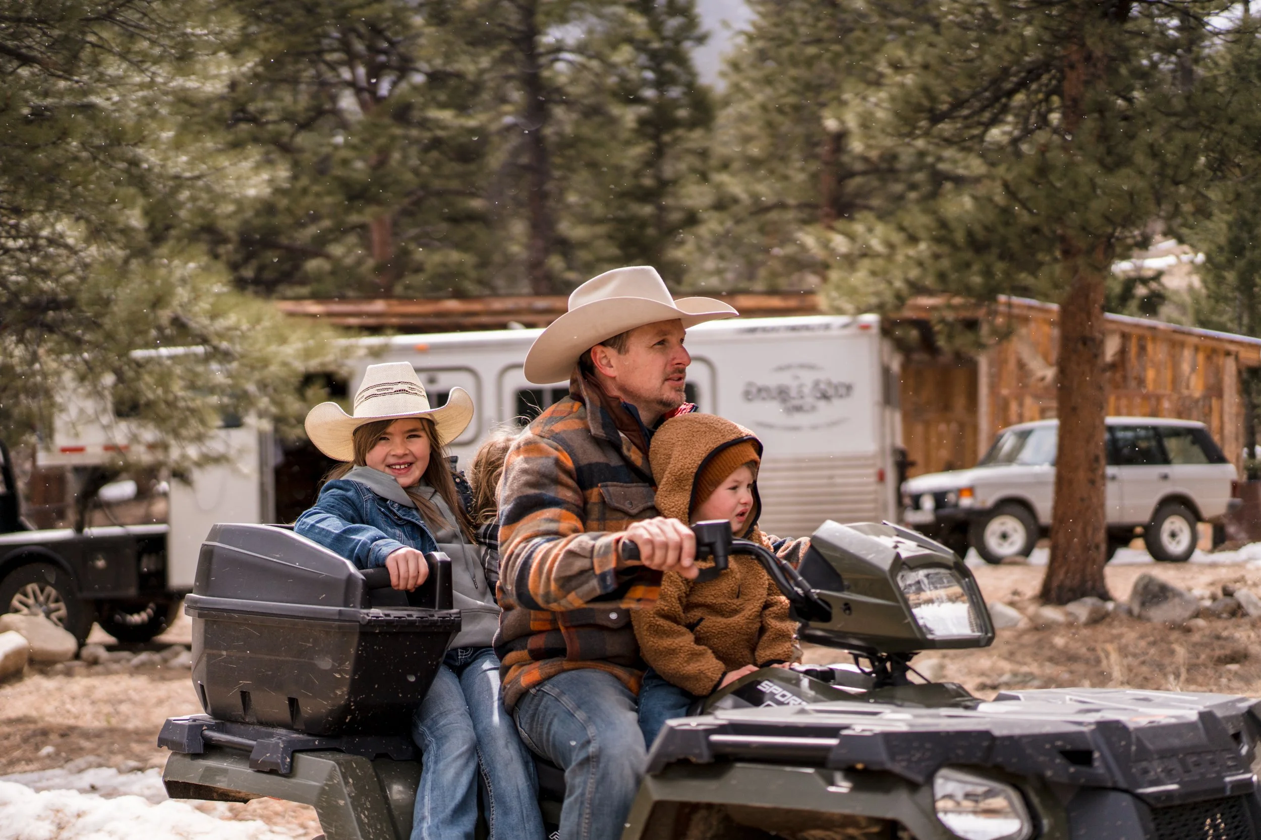 A man and three children on an ATV riding through a wooded area with snow on the ground. The man is wearing a cowboy hat and a plaid jacket, one girl is wearing a cowboy hat and smiling, and two young children are sitting in the back.
