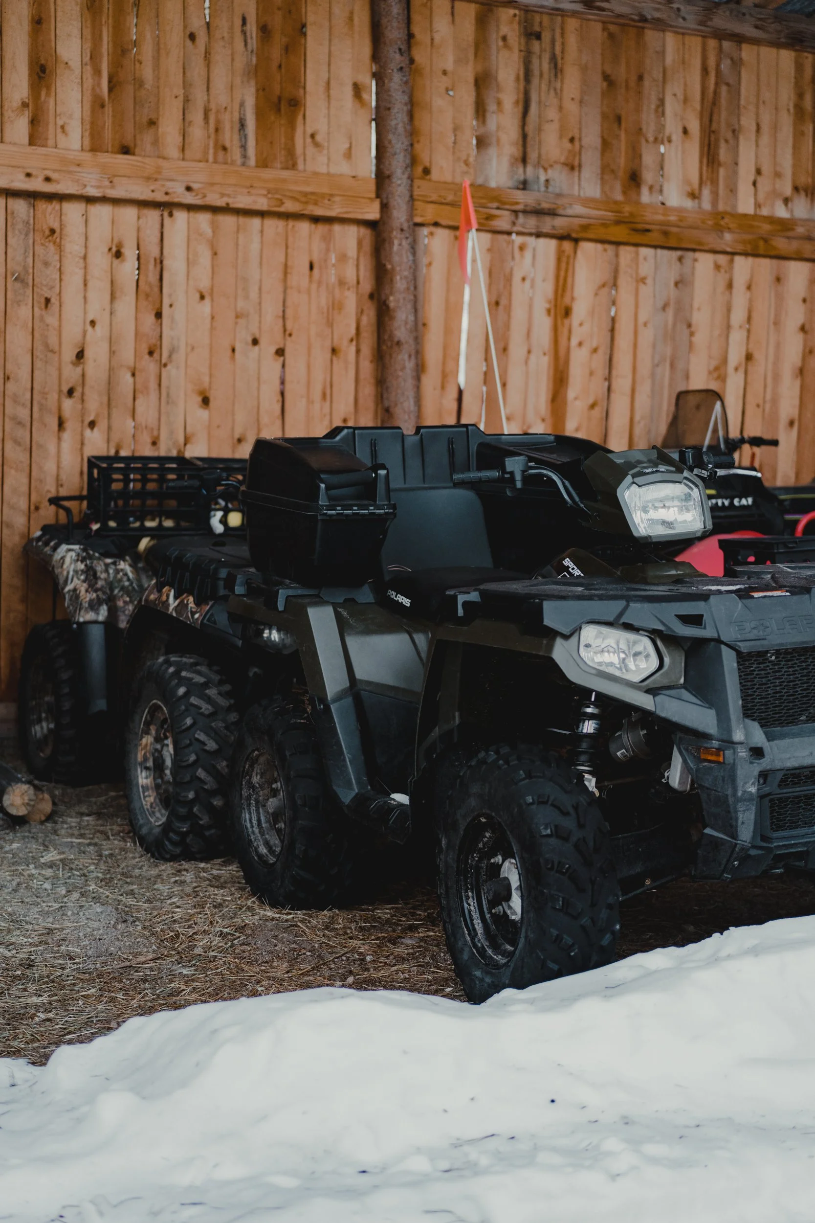 Six-wheeled black all-terrain vehicle (ATV) parked in a wooden shed with snow on the ground.