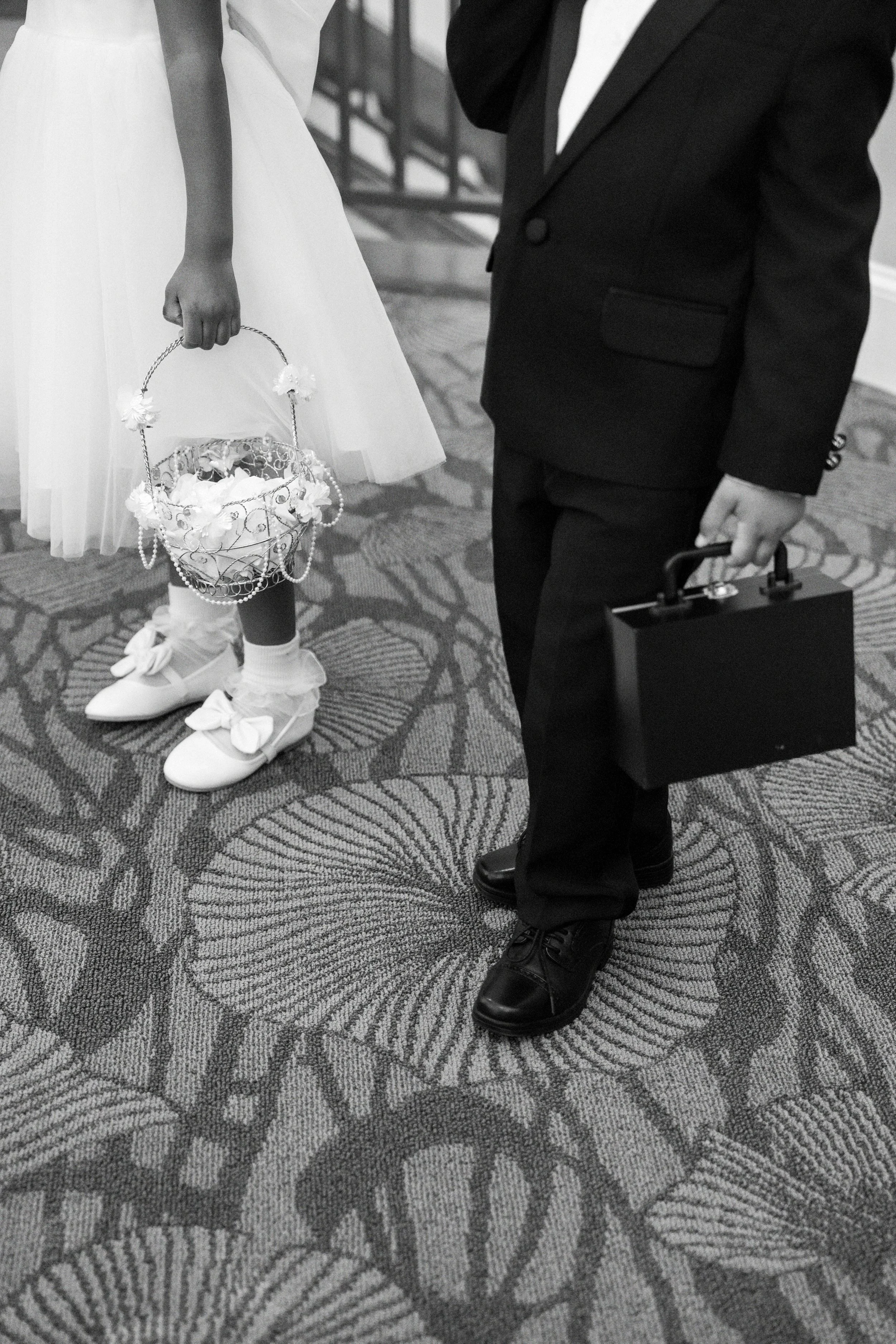 A black and white photo shows a young girl wearing a white dress with bow-tied shoes and holding a basket, standing next to a man in a dark suit holding a briefcase, on a patterned carpet.