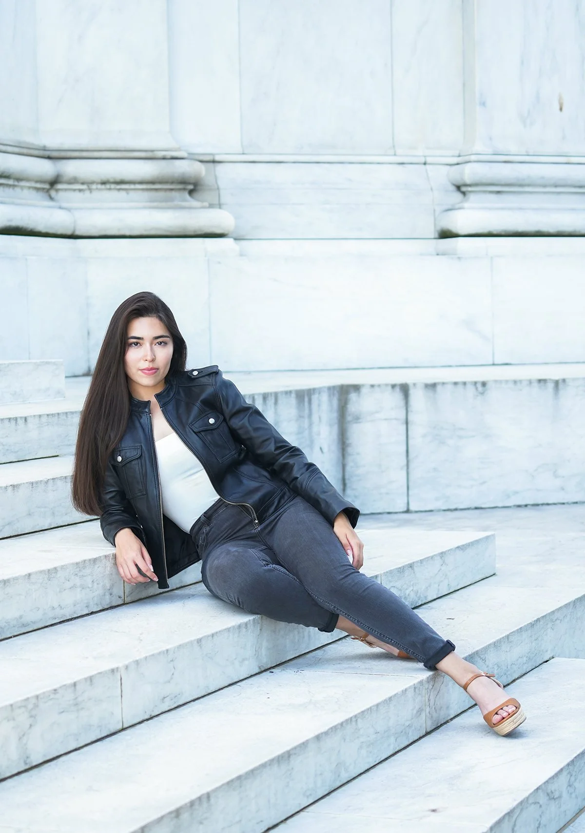 Young woman with long dark hair sitting on marble stairs outside, wearing a black leather jacket, white top, dark jeans, and beige wedge sandals.