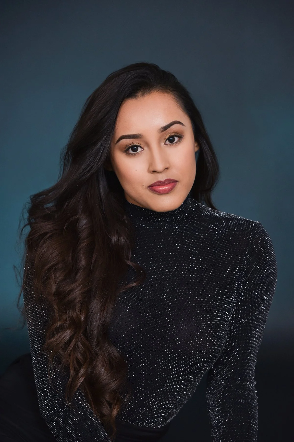 A woman with long, dark, wavy hair wearing a sparkly black turtleneck top.