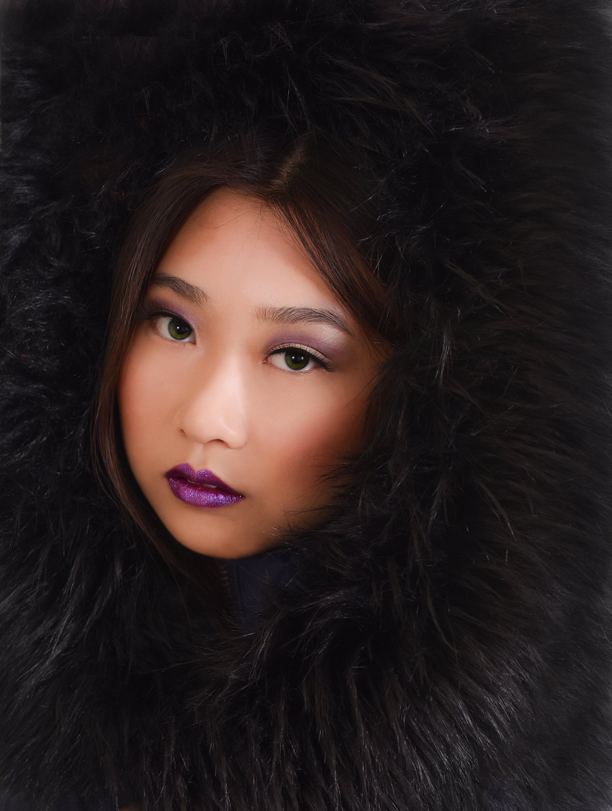 Close-up of a woman with green eyes and purple lipstick, wearing a black fur-lined hood or coat.