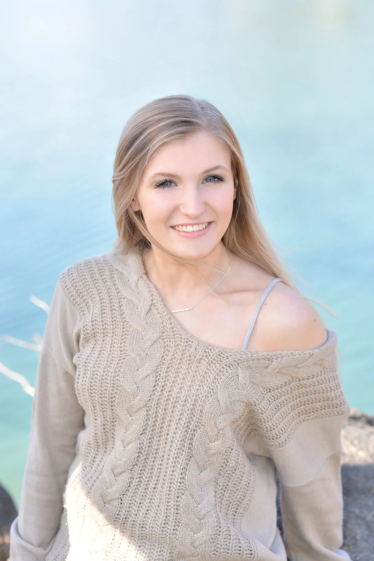 A young woman with blonde hair smiling outdoors near water, wearing a beige off-shoulder sweater and a silver necklace.
