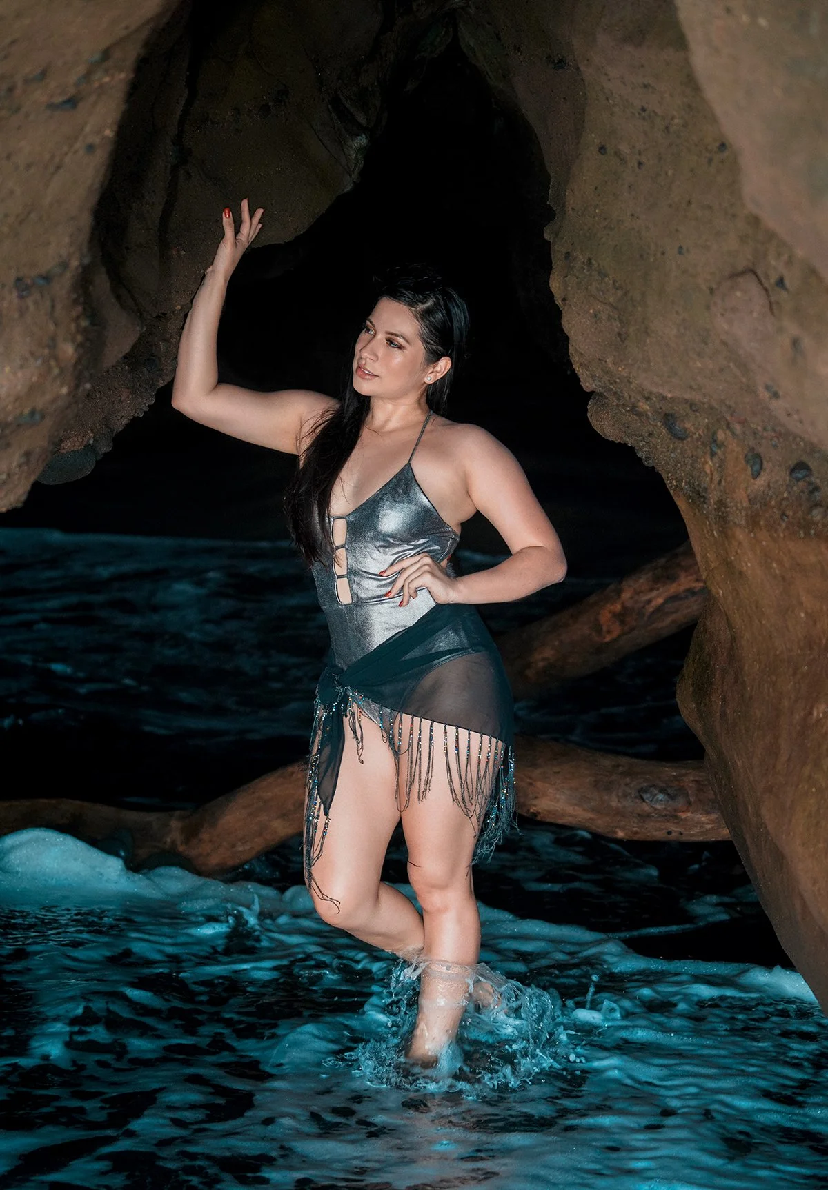 Woman in a metallic and black dress standing in water near a rocky cave entrance.