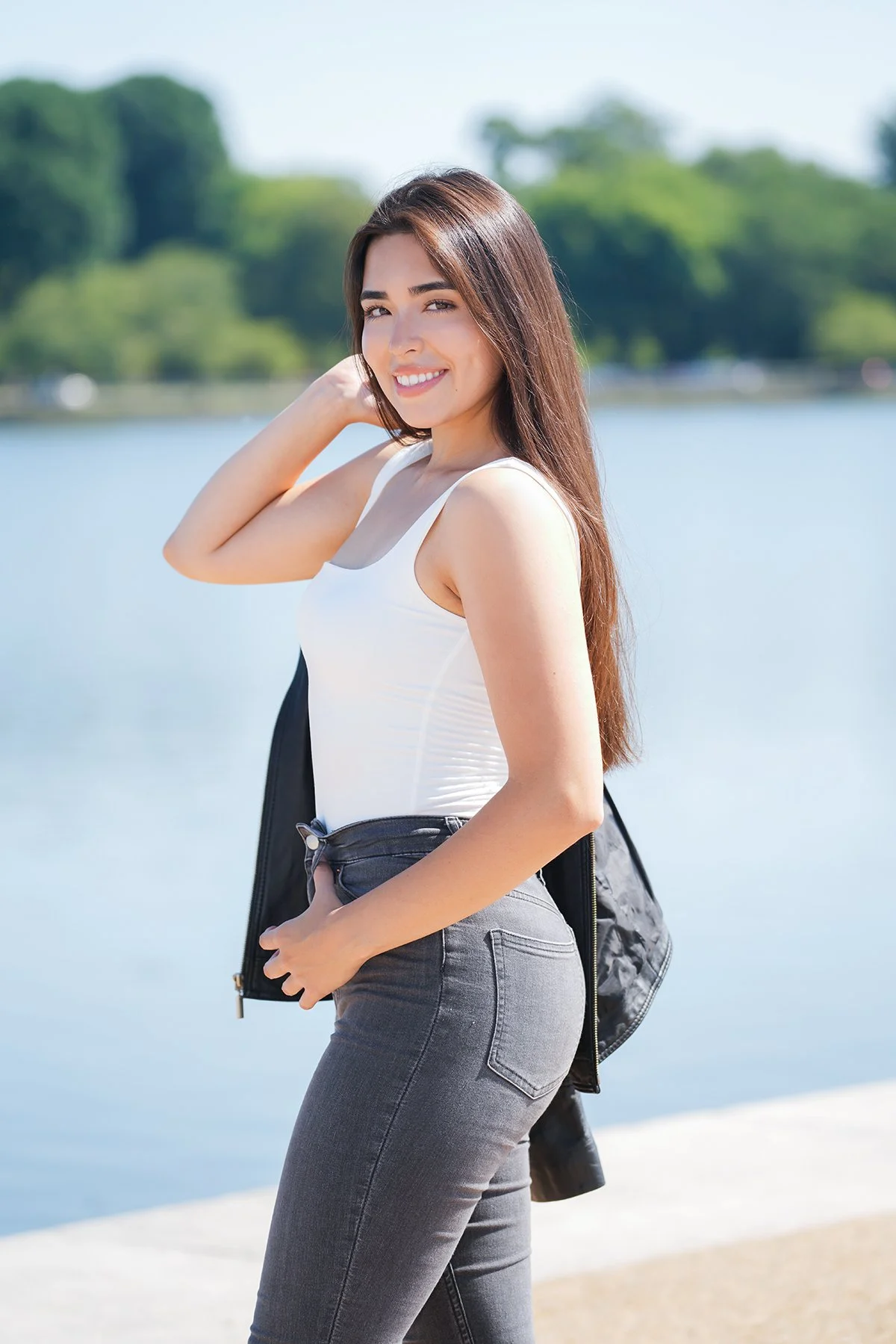 Young woman smiling and posing outdoors by a body of water, wearing a white tank top, gray jeans, and carrying a black jacket over her shoulder.