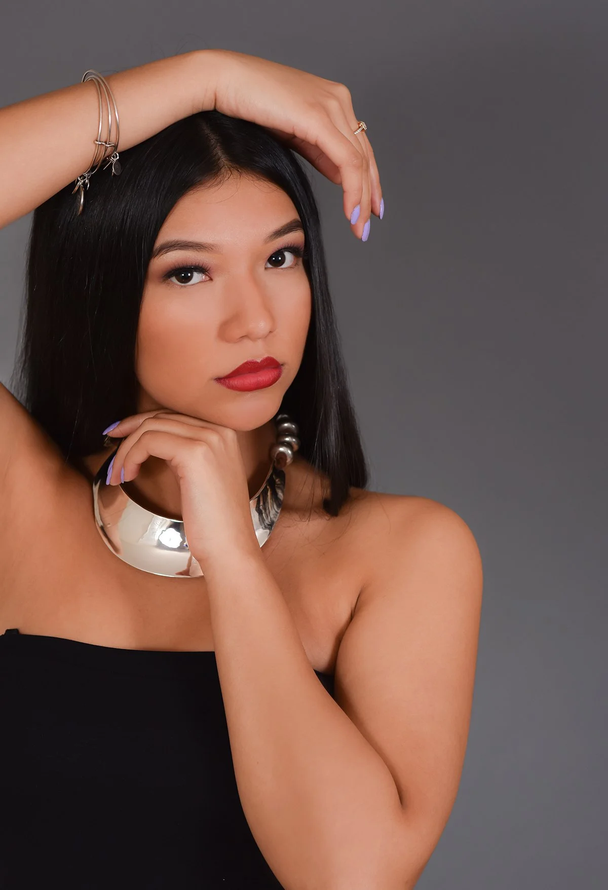 A woman with black hair and light skin posing against a gray background, wearing jewelry including a large metallic choker, earrings, and bracelets, with makeup featuring red lipstick and lavender nails.