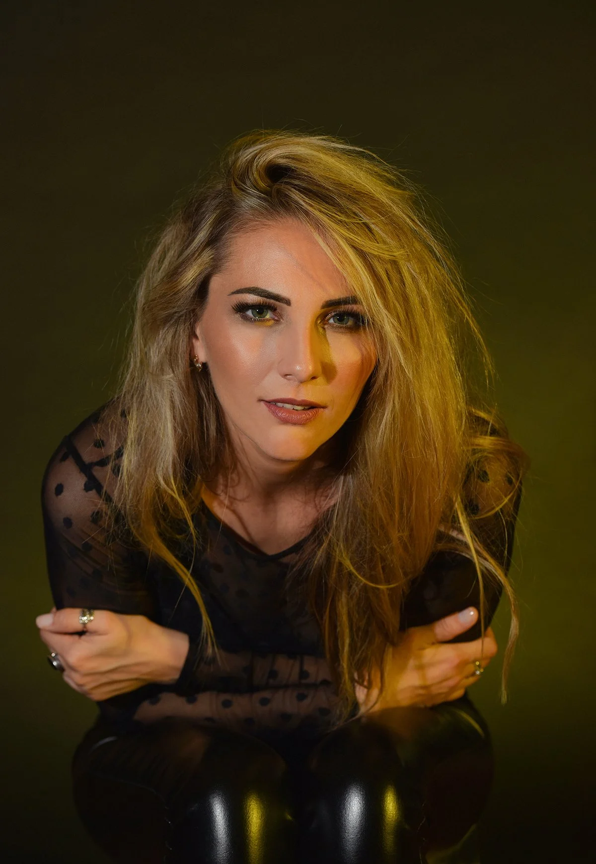 A woman with long, wavy blonde hair and green eyes is looking directly at the camera. She is wearing a black, sheer, polka-dot top and has rings on her fingers. Her arms are crossed in front of her, and she has a confident expression.