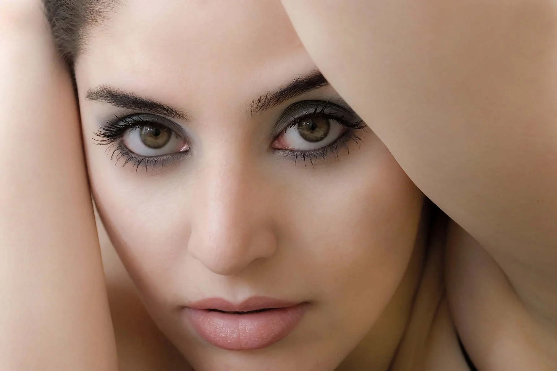 Close-up of a woman's face with brown eyes, dark makeup, and relaxed expression, partially framed by her arms.