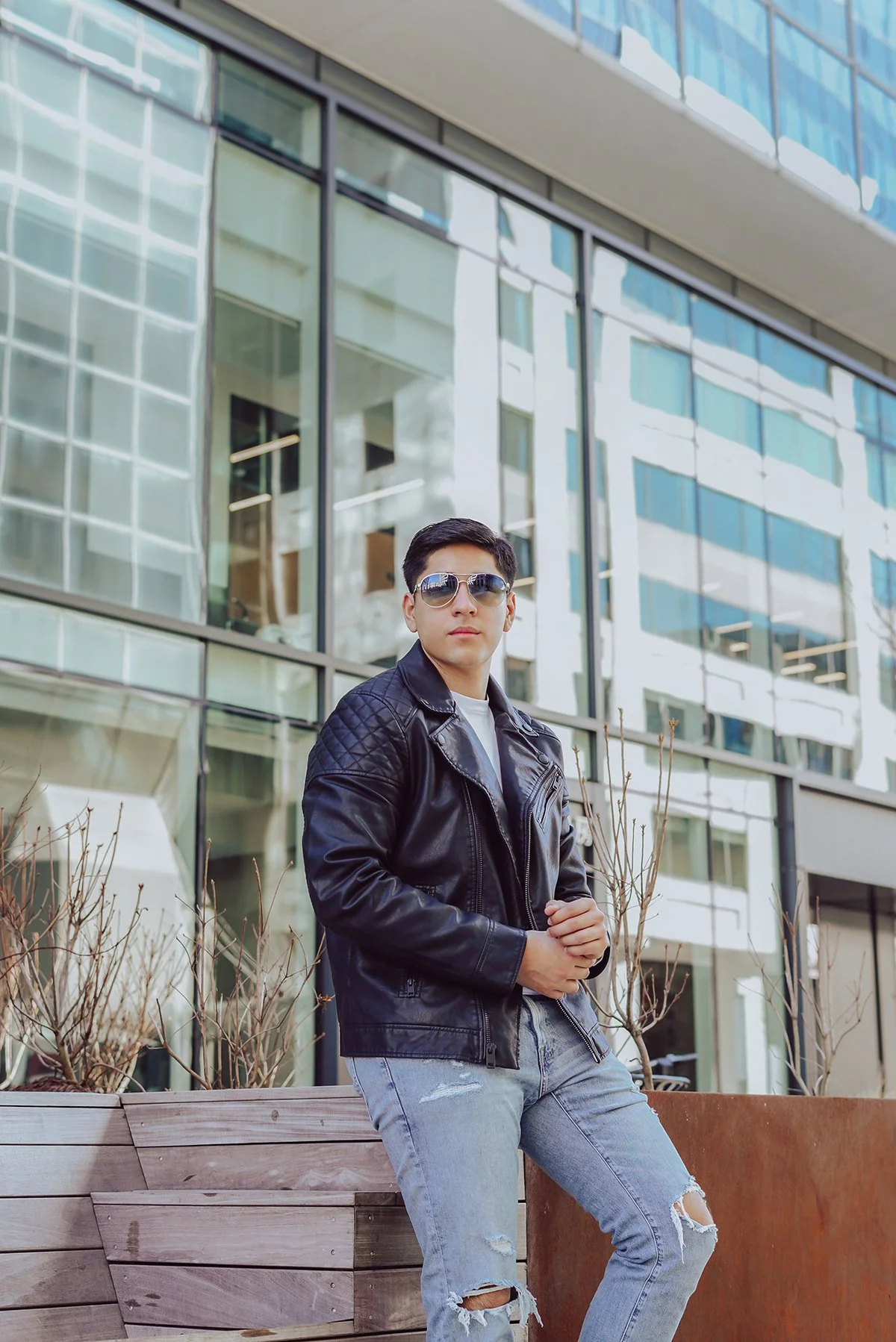 Young man wearing sunglasses, a black leather jacket, and ripped jeans standing outdoors in front of a modern glass building.