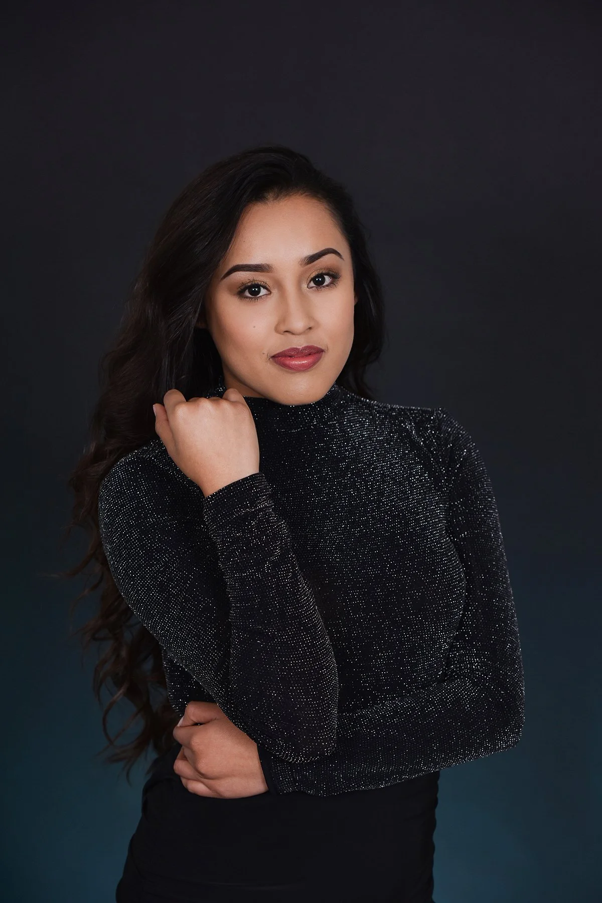 A young woman with long, dark hair, wearing a black dress with shimmering details, poses against a dark background, resting her chin on her hand.